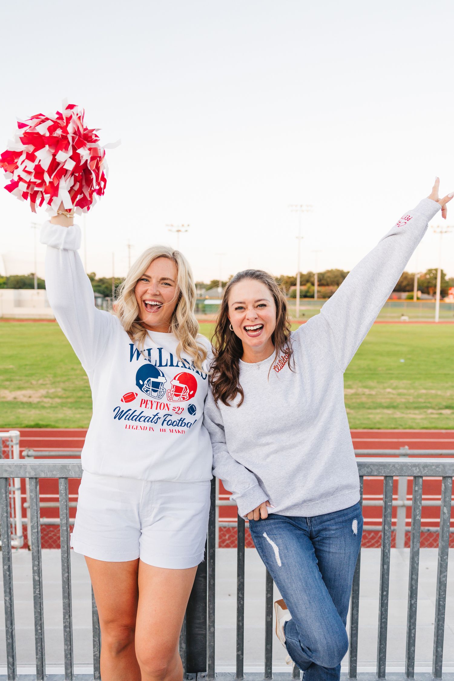 Two women cheering at football stadium wearing team sweatshirts, one holding red and white pom poms.