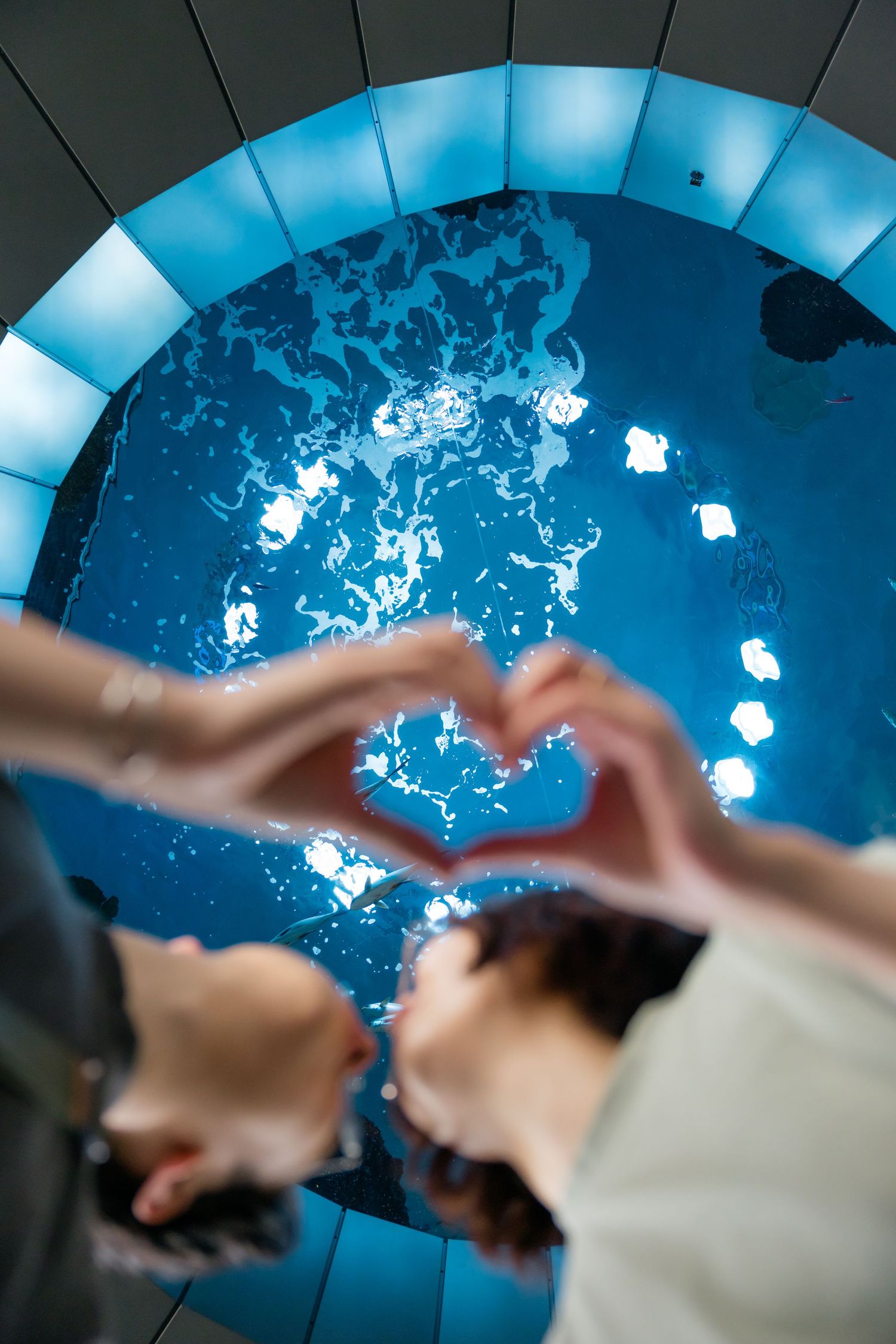 Two people forming a heart shape with their hands over a glowing blue circular water feature.