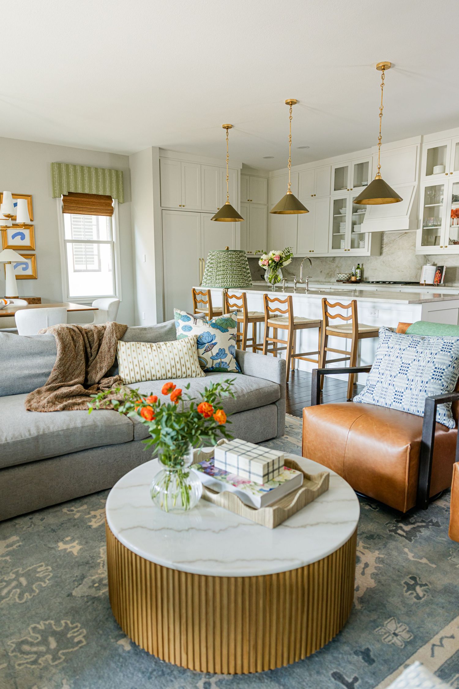 Open-concept living room with gold fluted coffee table, marble top, leather chair, and kitchen with brass pendant lights.