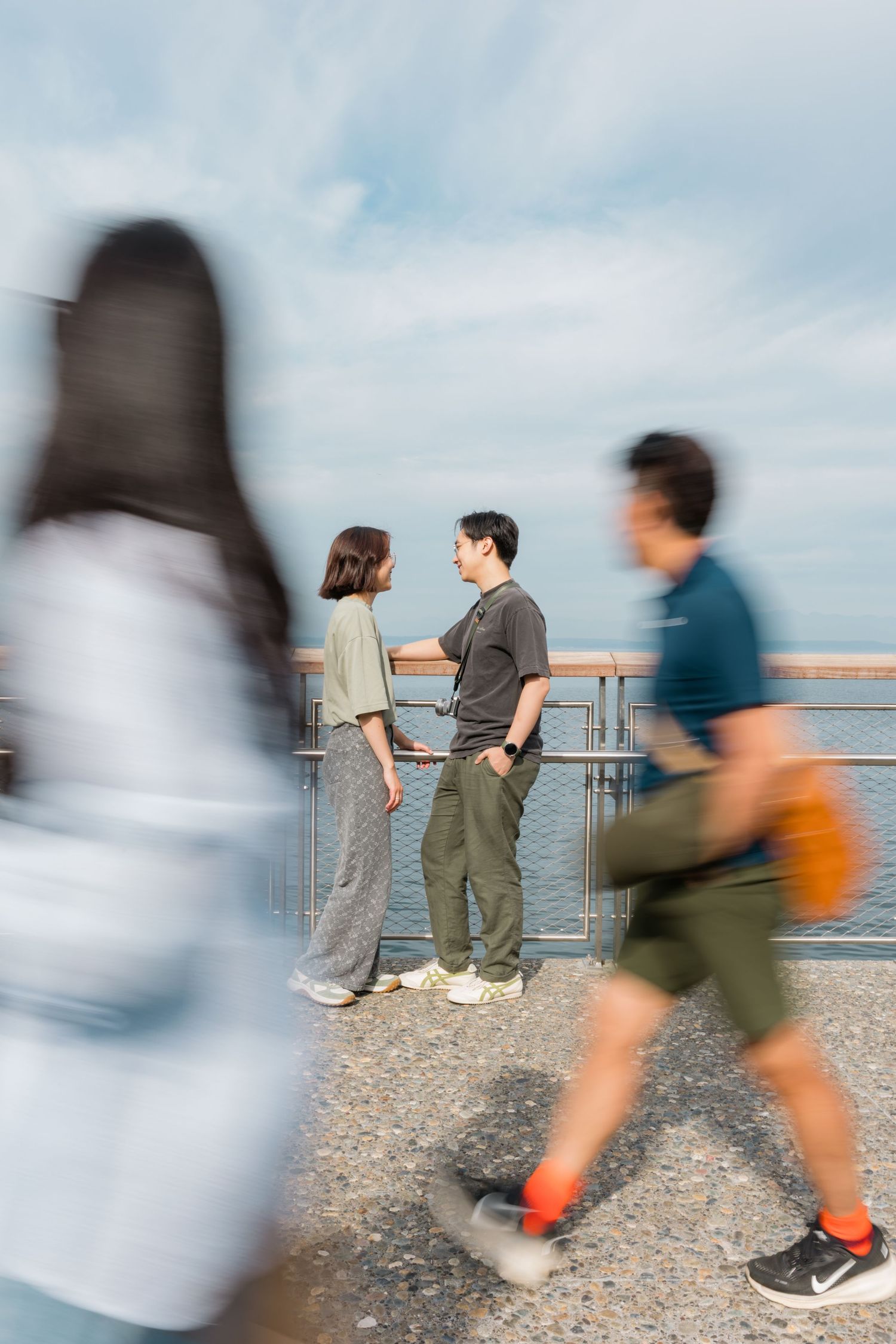 Two people conversing by a waterfront railing while blurred pedestrians pass in the foreground on a cloudy day.