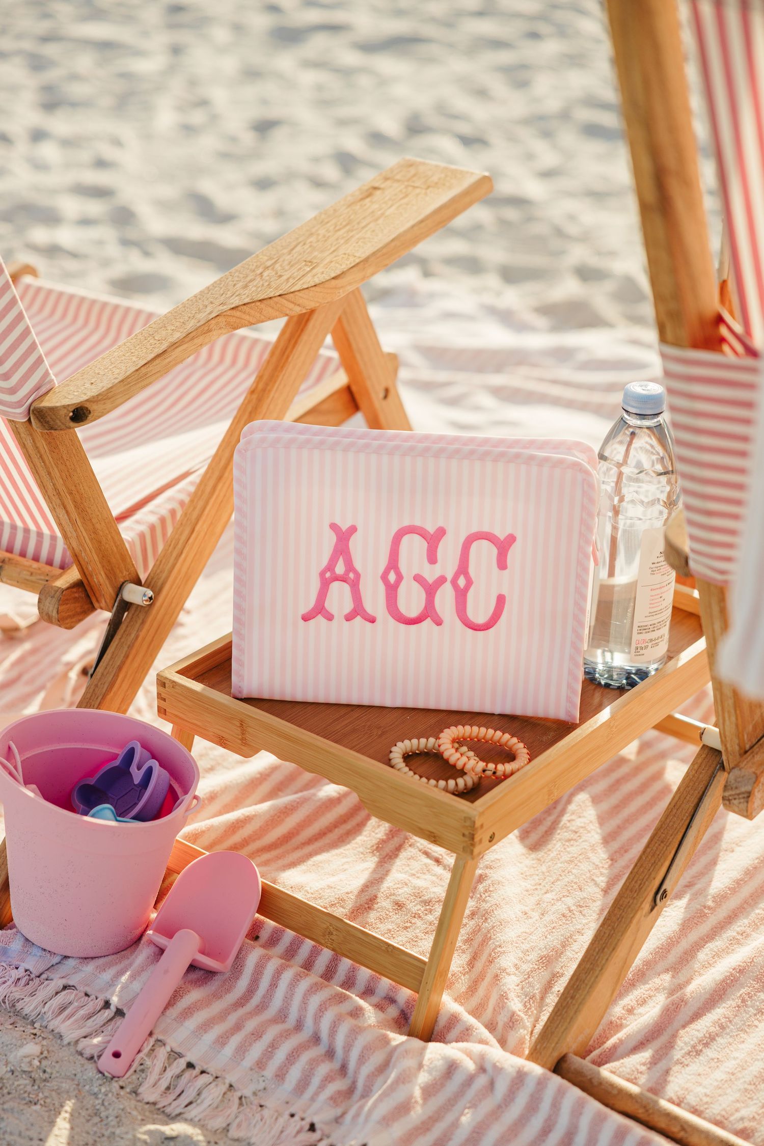 Monogrammed AGC pink striped pouch on wooden beach tray with water bottle and pearl bracelet.