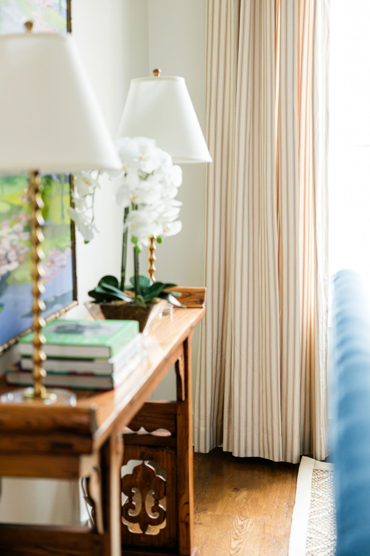 Wooden console table with gold lamps, orchid plant, and green books beside striped curtains in a bright room.
