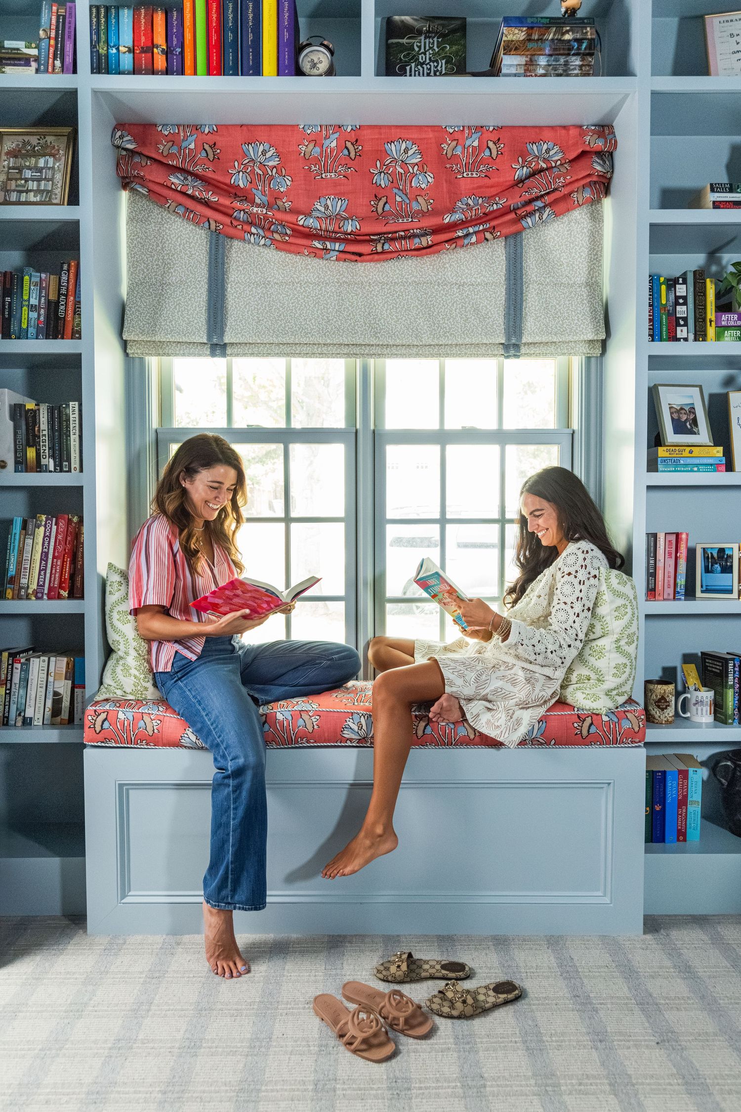 Two women reading books on a built-in window seat surrounded by blue bookshelves in a cozy home library.