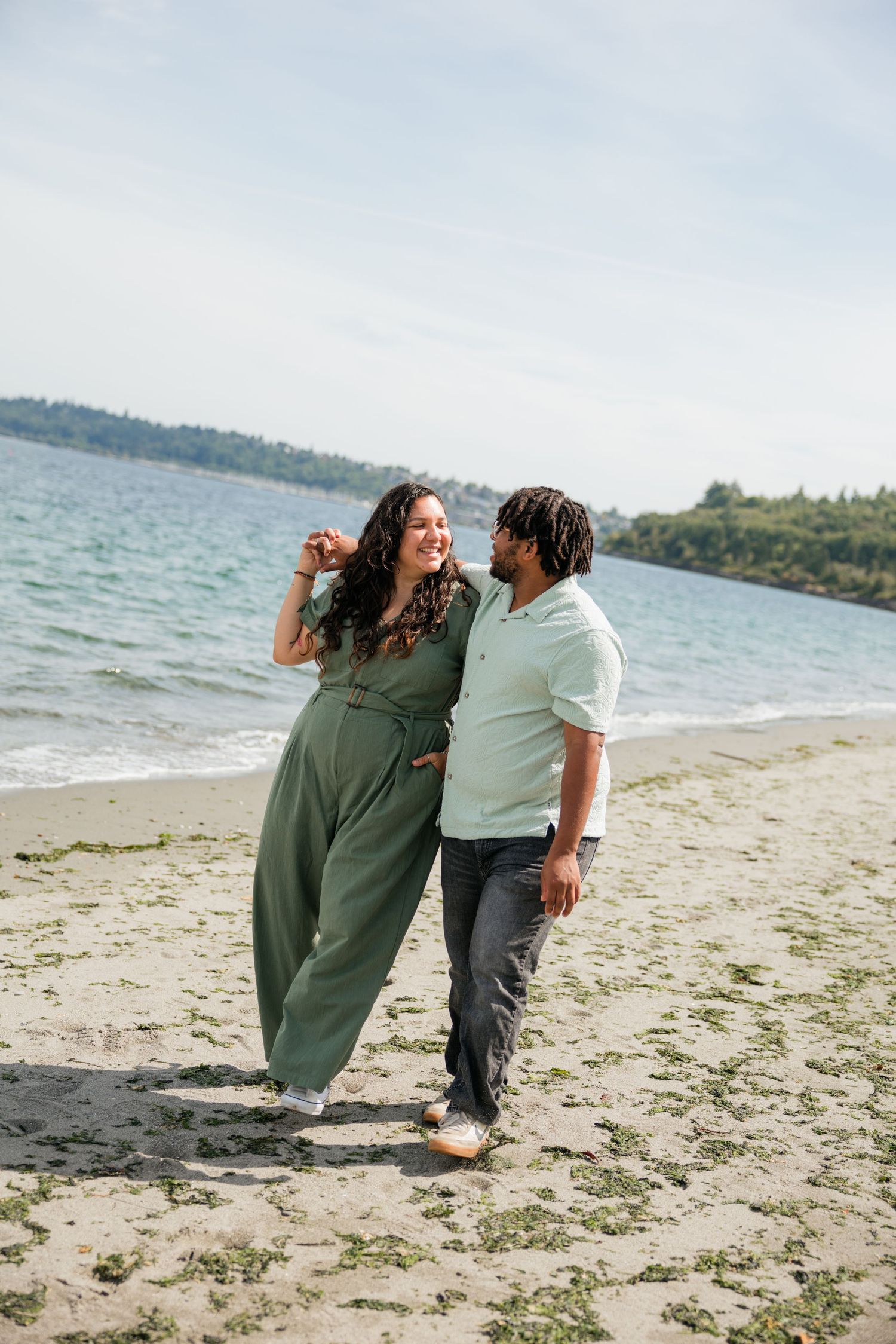 Laughing couple in coordinating green and mint outfits stroll hand-in-hand along a scenic beach.