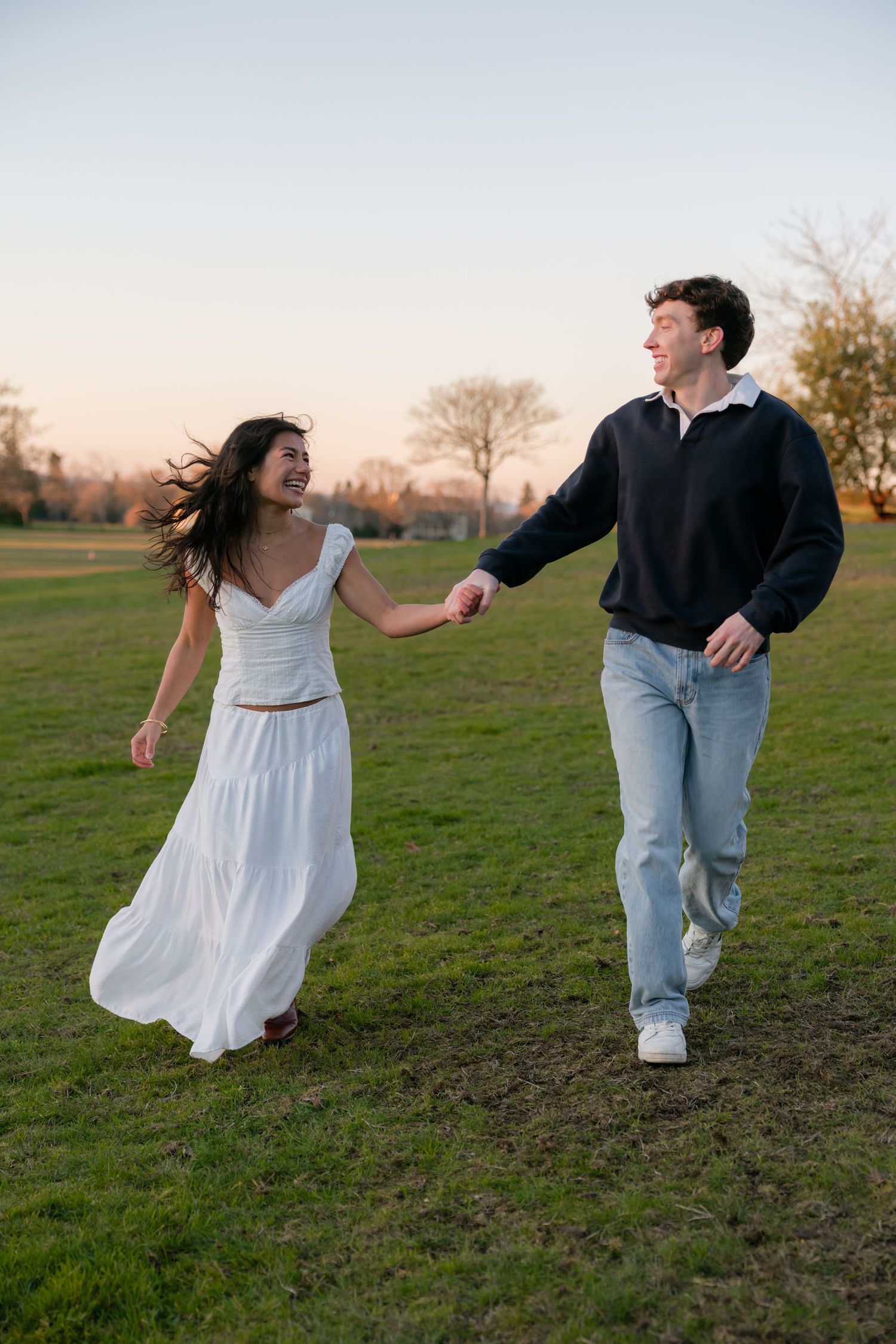 A joyful couple holds hands while running through a green park field at golden hour sunset.