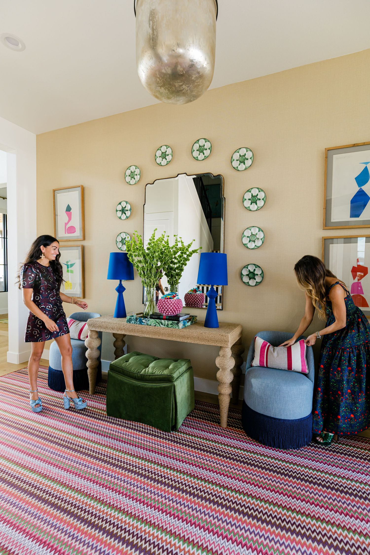 Two women arranging colorful decor with blue lamps and ottomans in bright entryway with chevron rug.