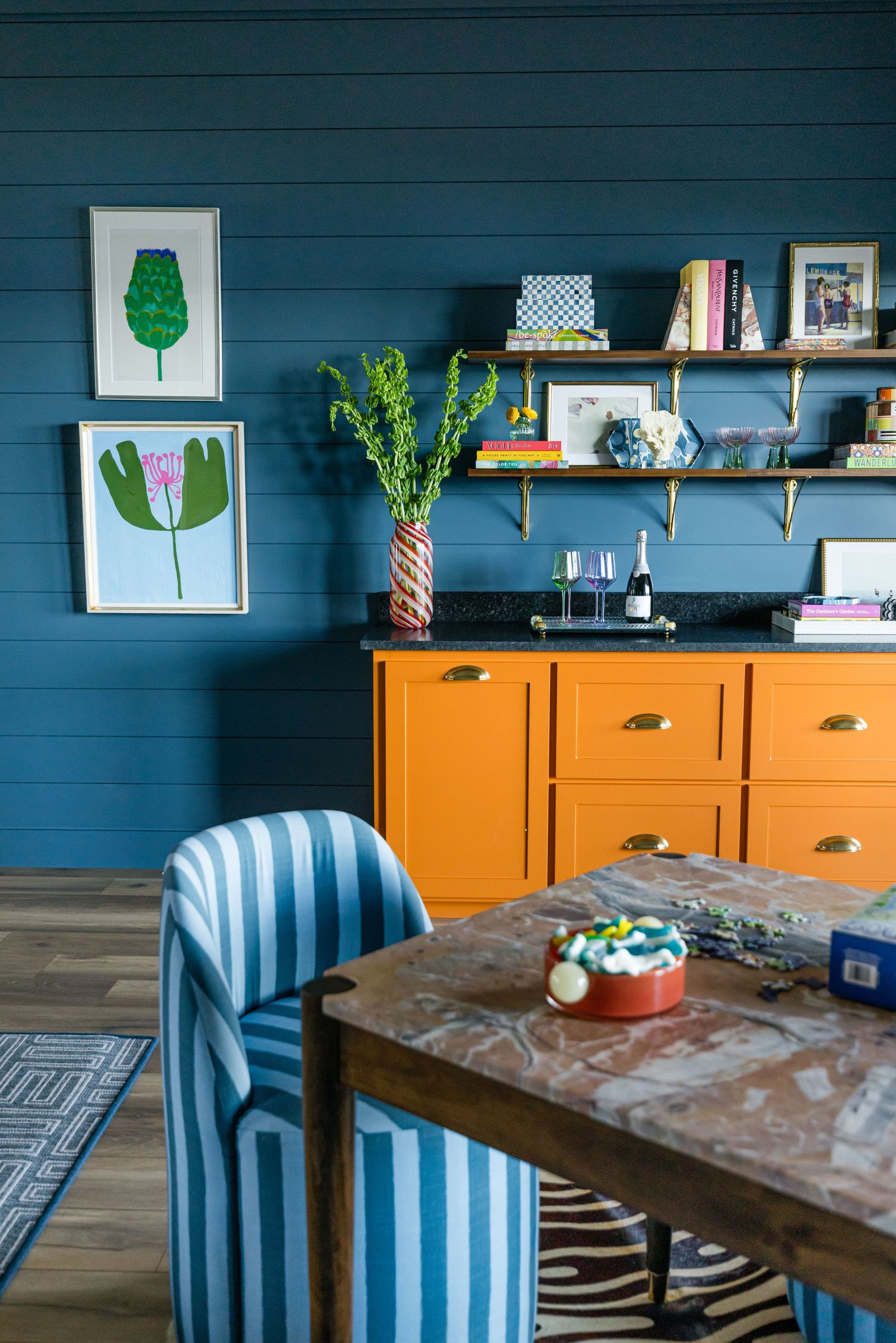Bold blue shiplap dining room with orange cabinet, striped chairs, botanical art, and brass open shelving.
