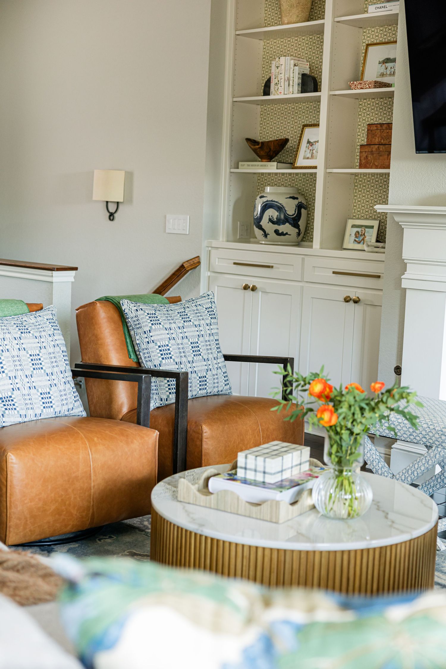 Tan leather chairs with blue patterned pillows beside a gold and marble round coffee table with orange flowers.