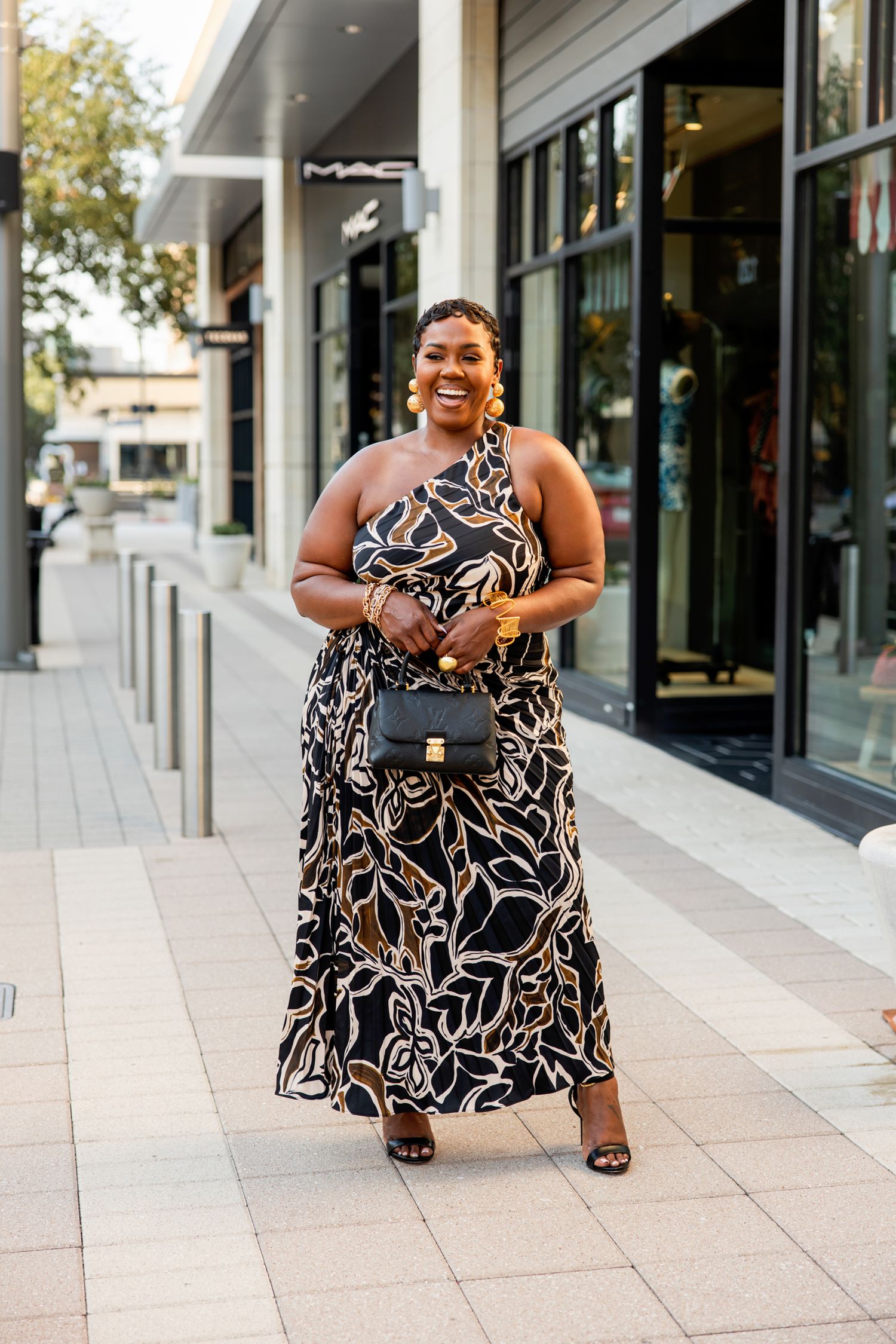 Joyful woman in bold floral one-shoulder maxi dress walking past shops while carrying a black bag.