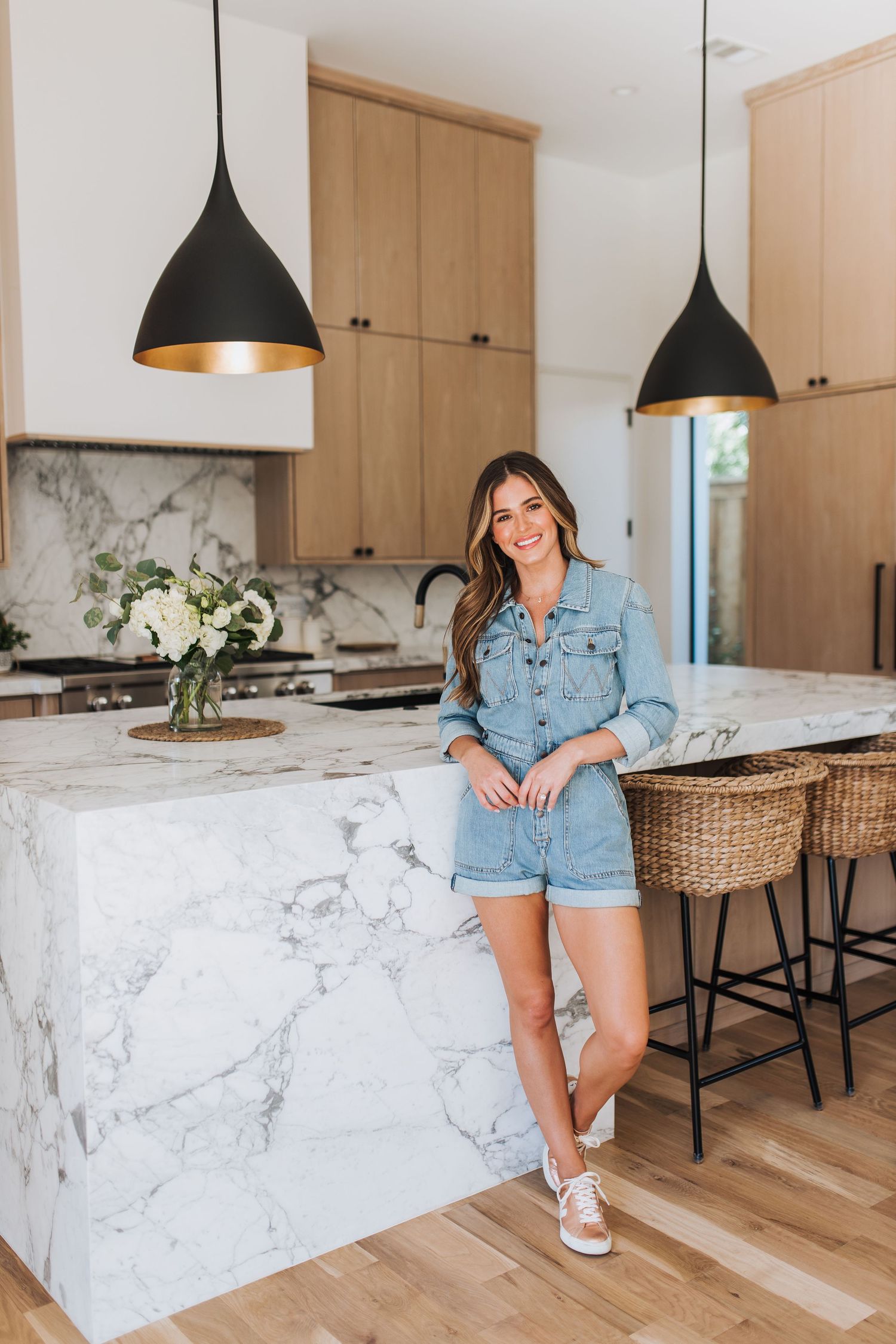 Smiling woman in denim romper posing beside marble kitchen island with black gold pendant lights overhead.