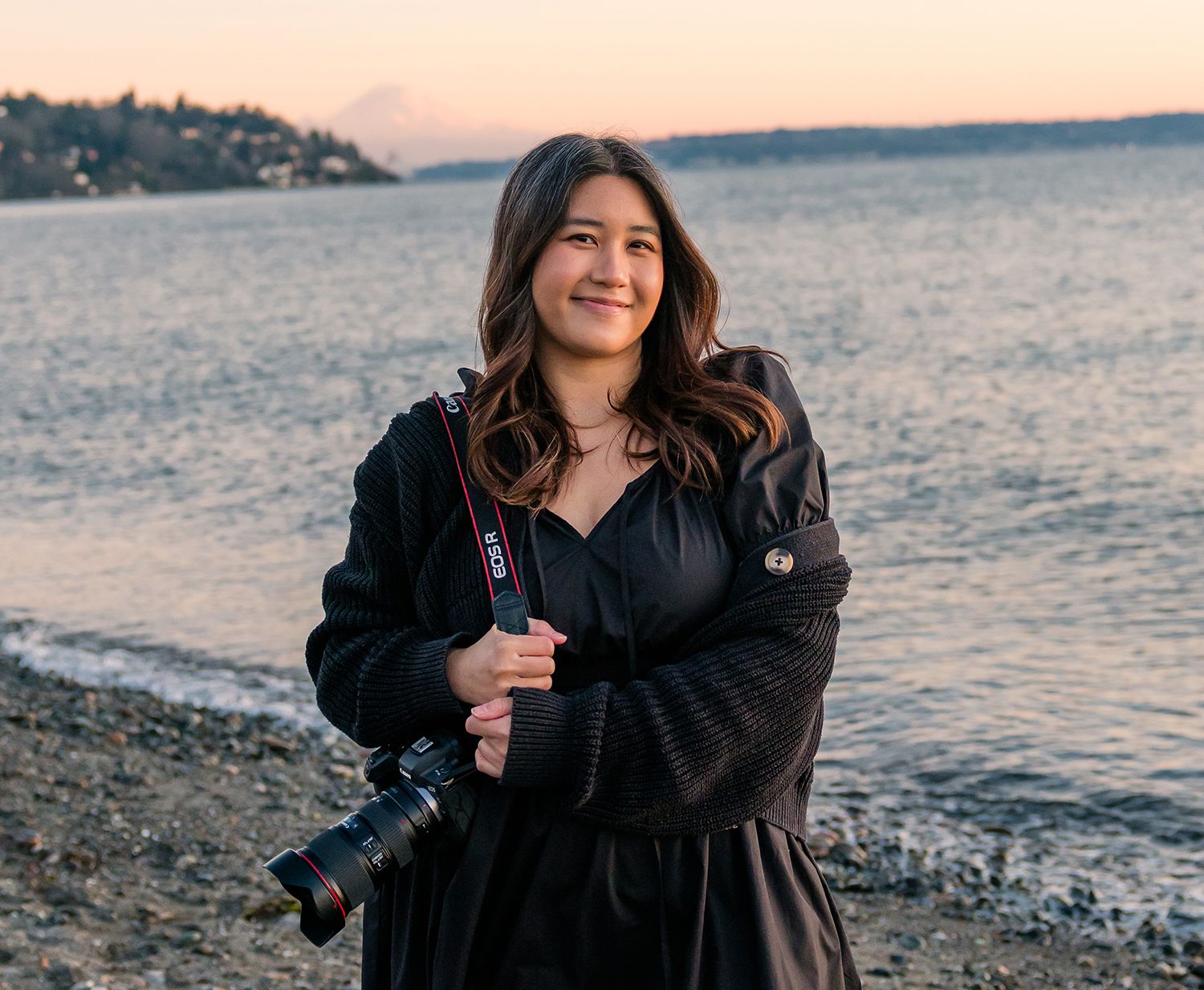 Smiling young woman holding a Canon camera stands on a pebble beach at sunset with calm water behind her.