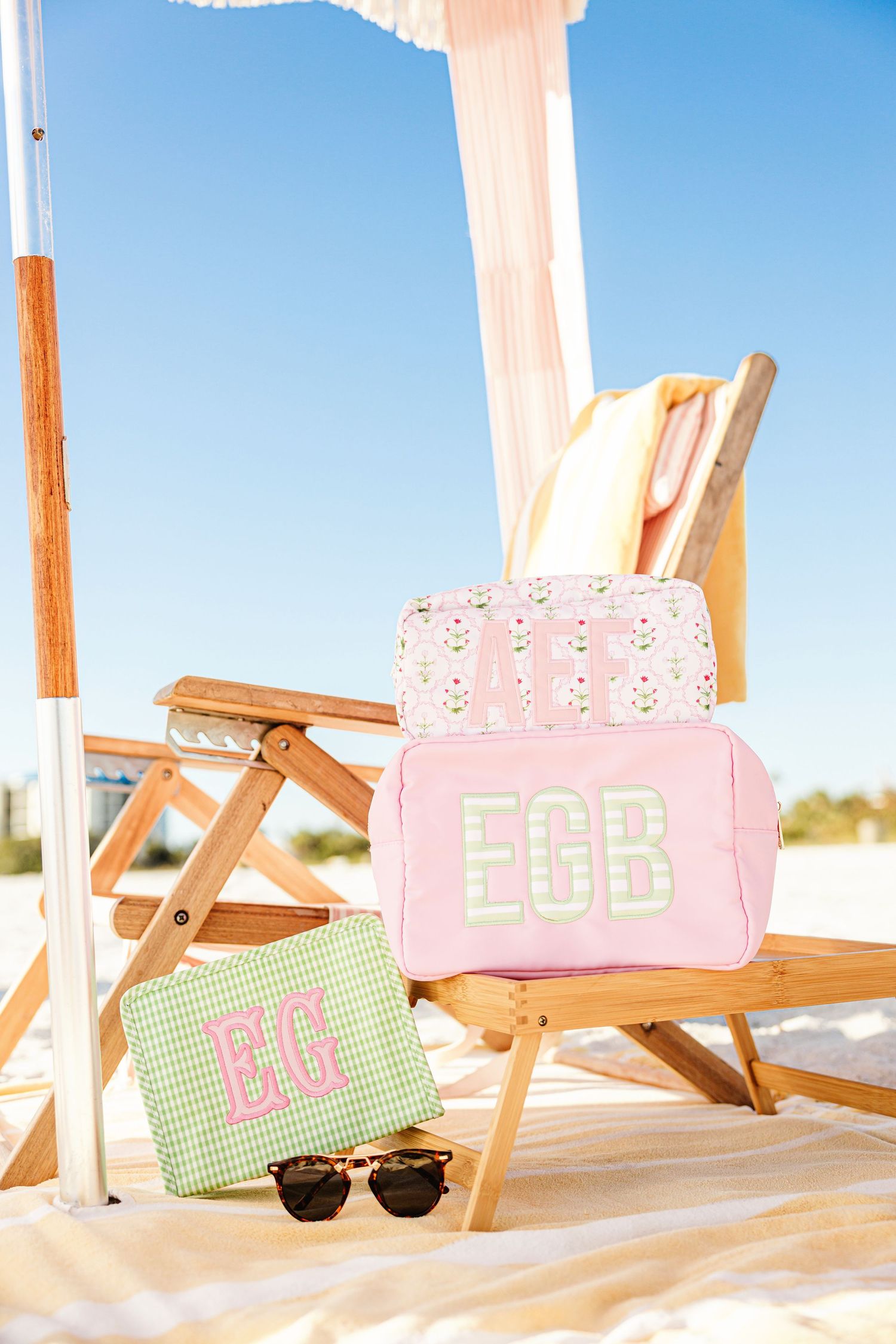 Stacked monogrammed pouches EGB pink and EG green gingham on wooden beach chair in sand.