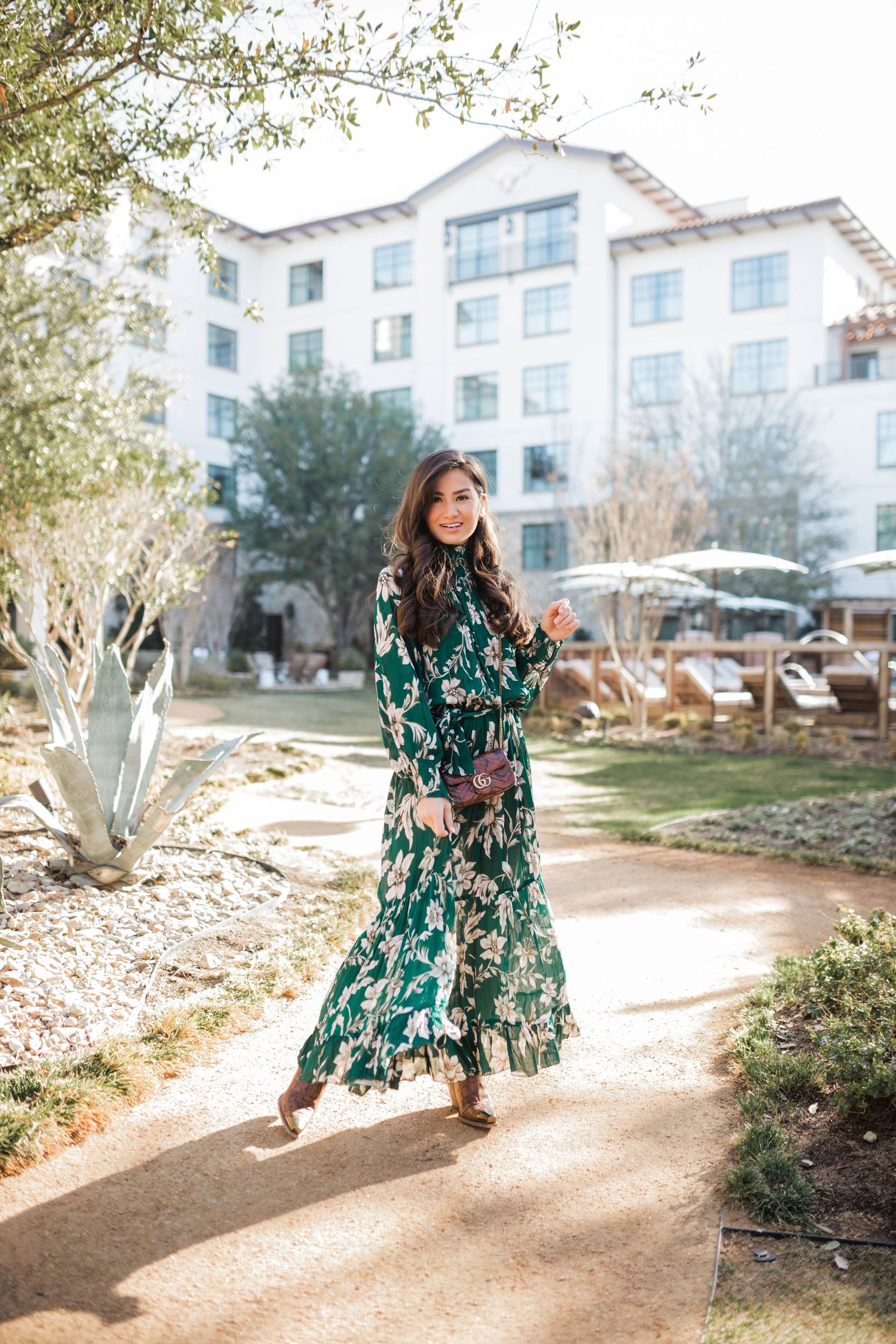Woman in green floral maxi dress poses on garden path outside luxury hotel on sunny day.