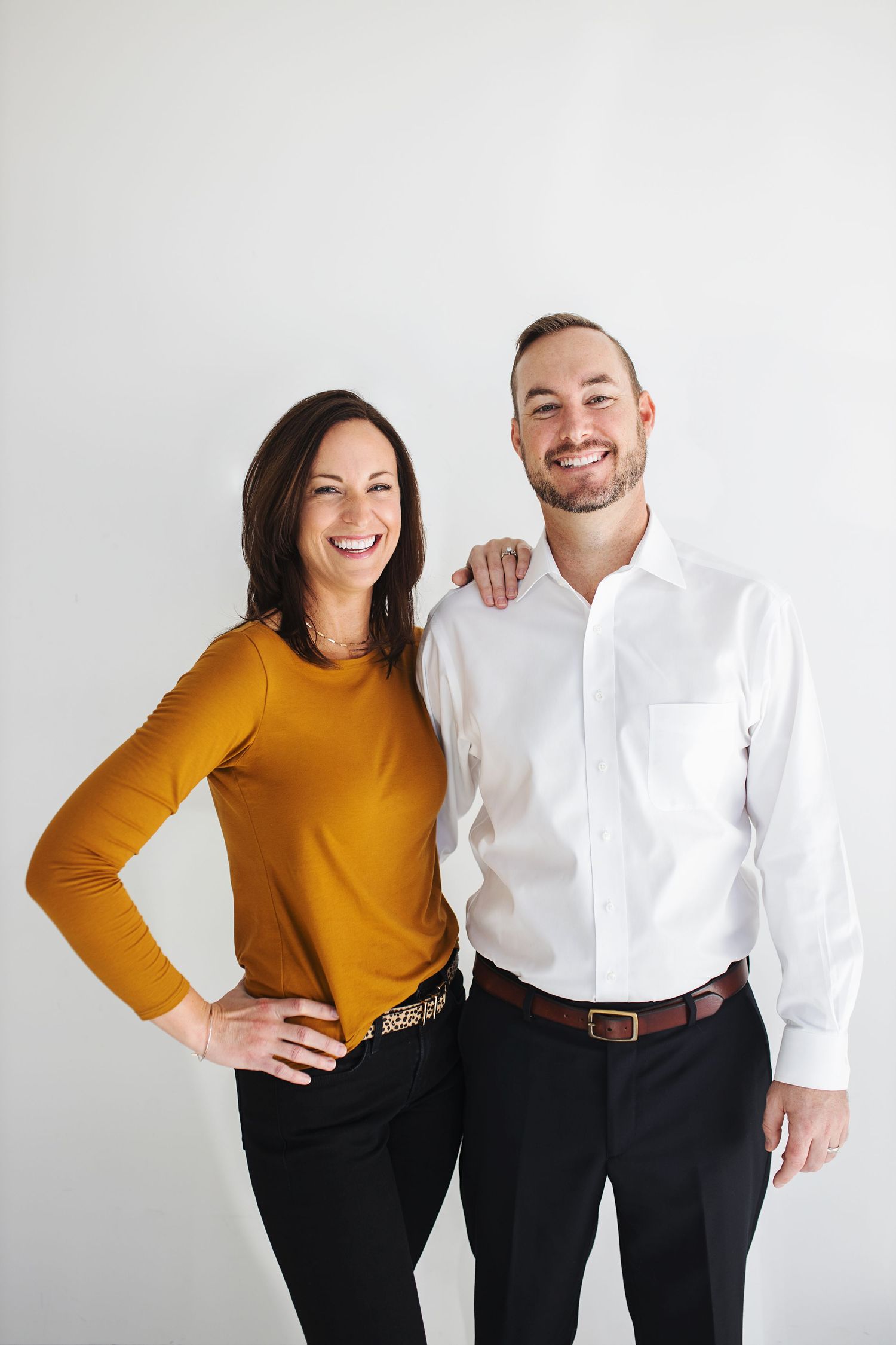 A smiling man and woman pose together against a white background, she in a mustard top, he in a white dress shirt.