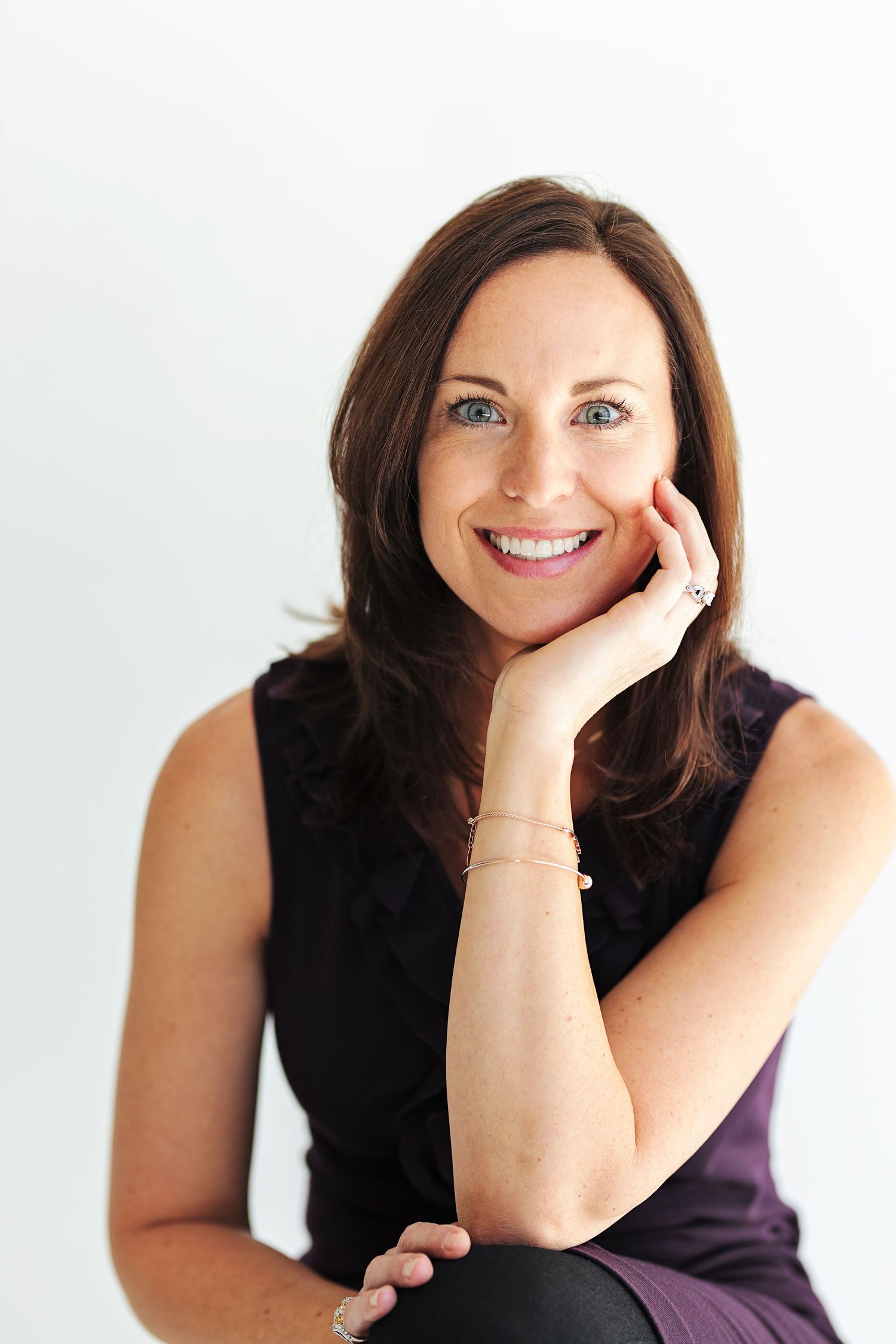 Smiling brunette woman in black sleeveless top resting chin on hand, wearing gold bracelets, against white background.