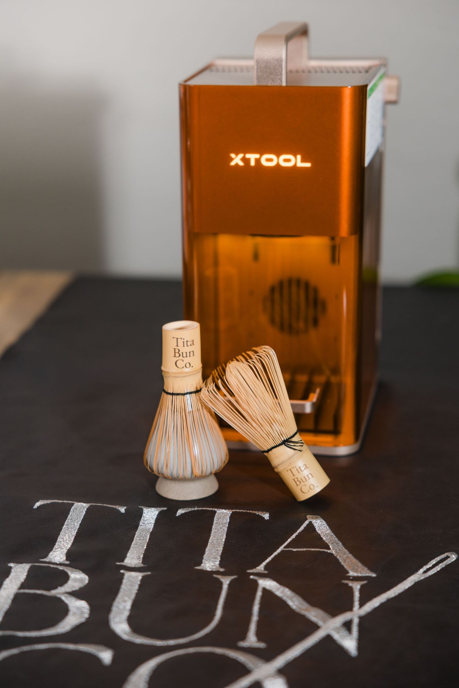 Two bamboo matcha whisks with Tita Bun Co. branding displayed in front of an xTool laser engraver.