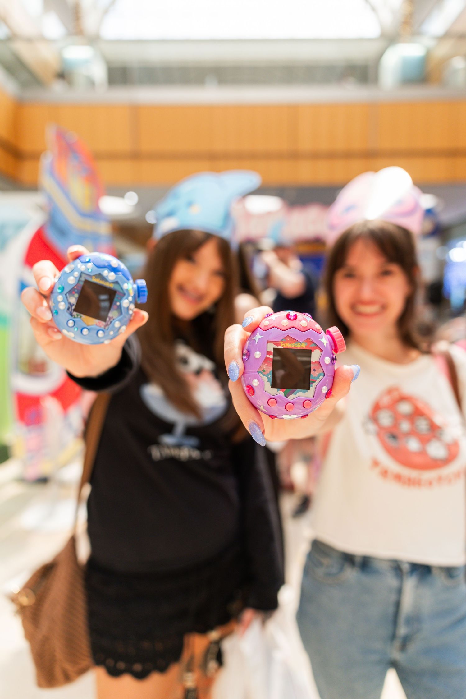 Two women hold up blue and pink Tamagotchi devices toward the camera at a mall event.