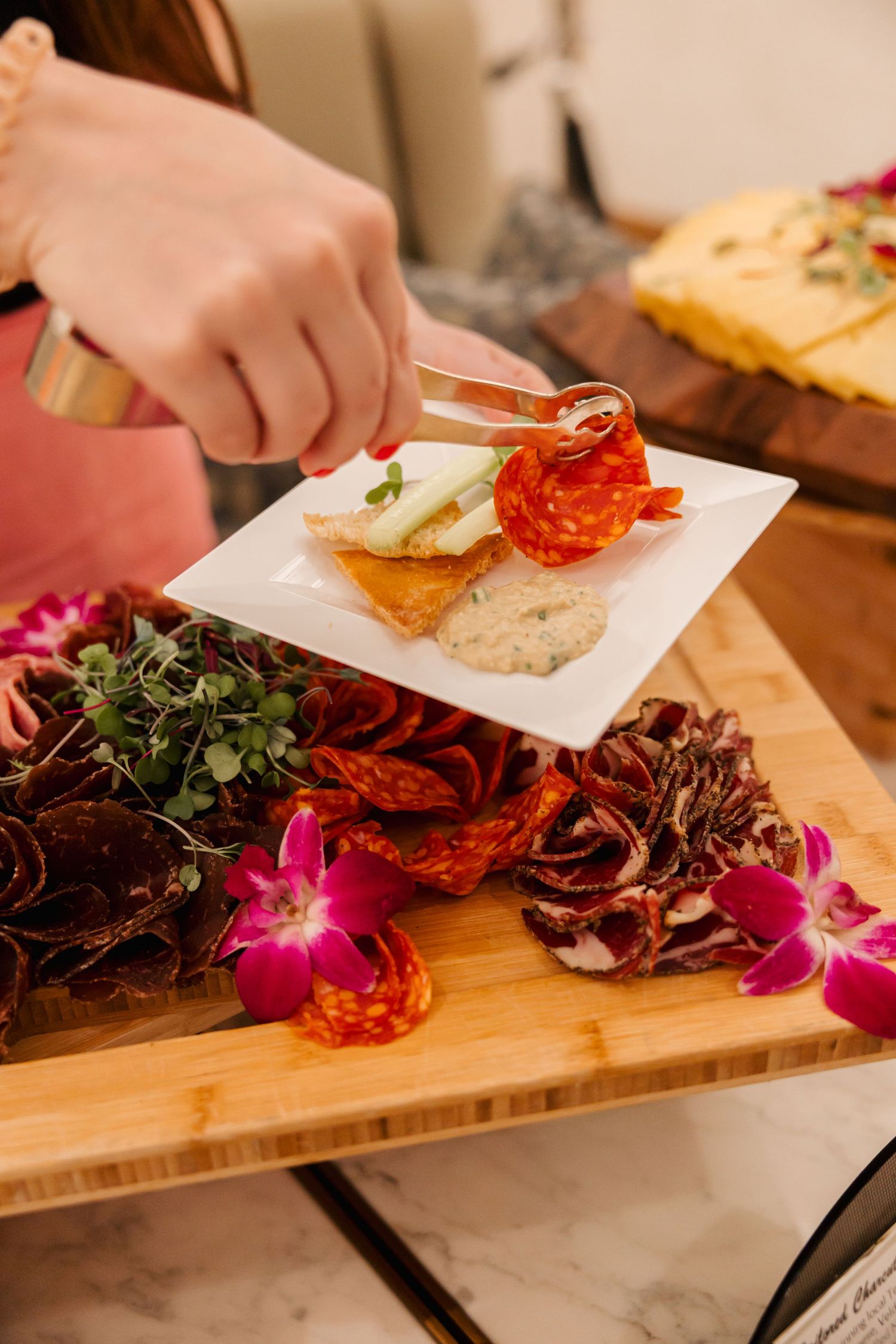 Hand using tongs to serve charcuterie and appetizers on a decorative wooden board.