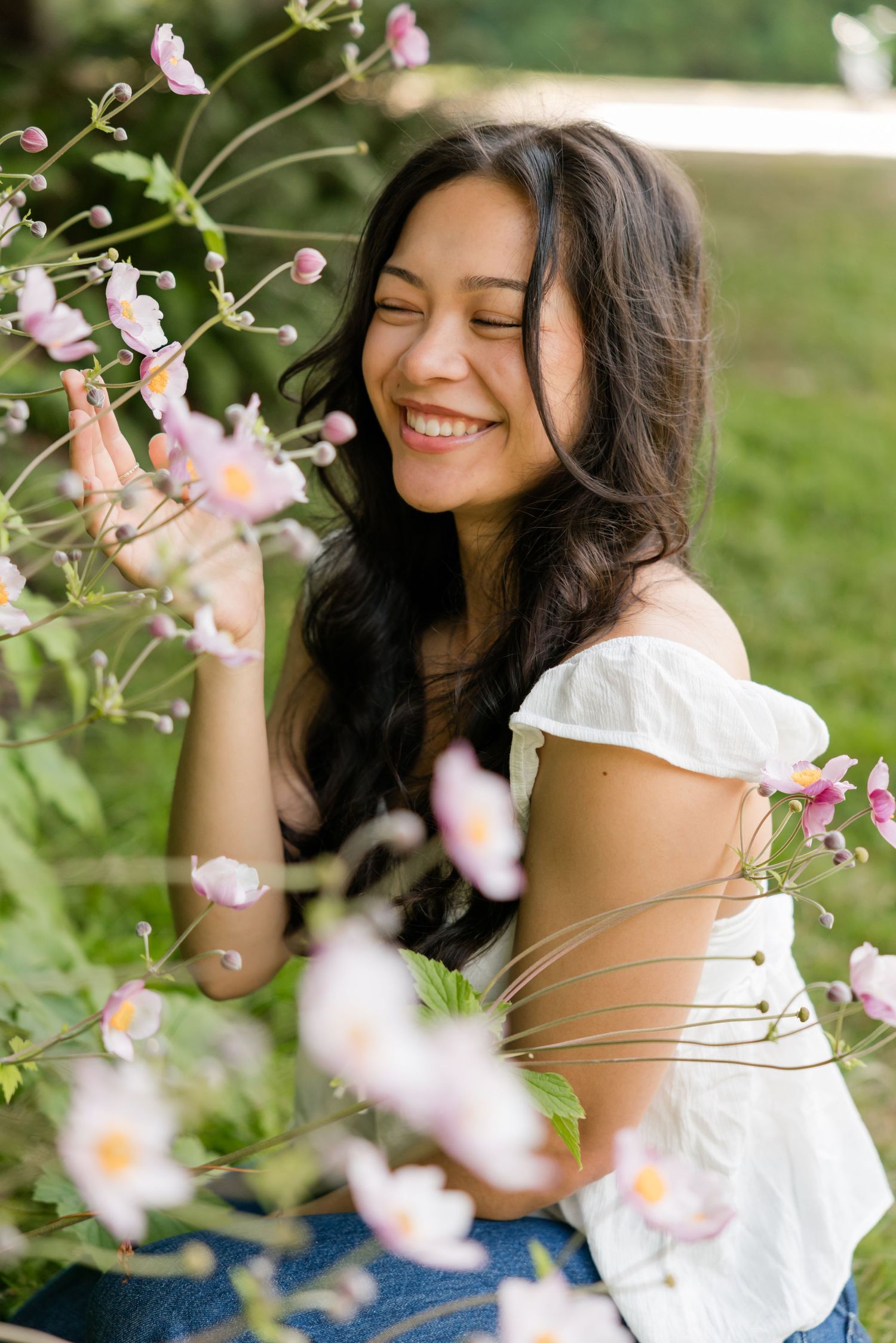 Happy young woman smiles with eyes closed among delicate pink flowers in a sunlit garden setting.