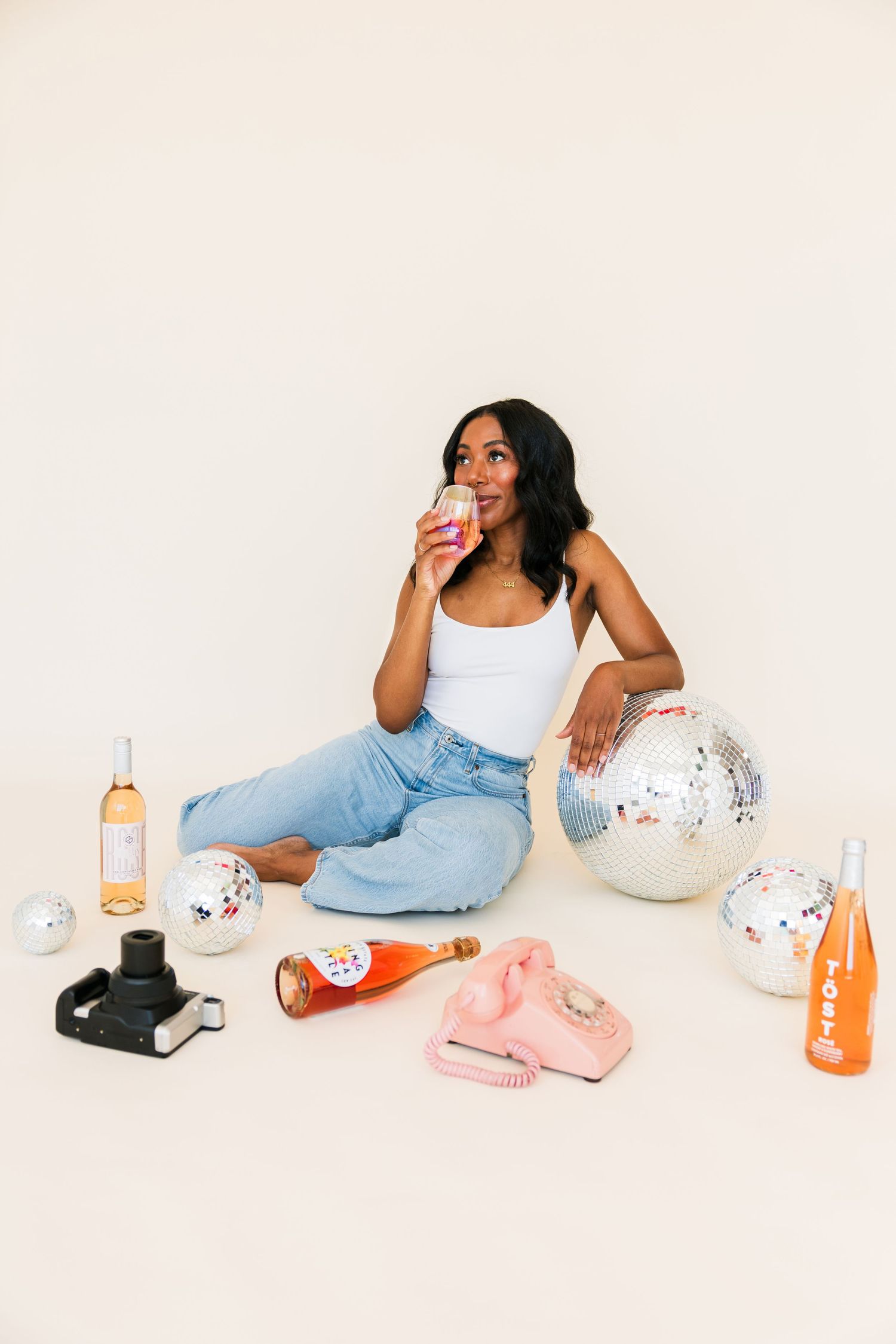Woman in white tank top sipping drink while seated among disco balls, wine bottles, and vintage pink phone on floor.