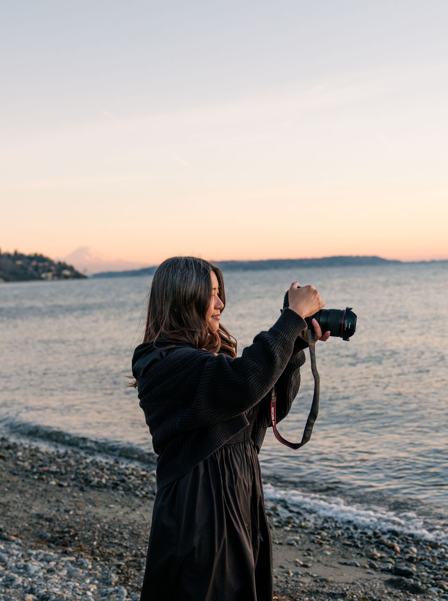 Smiling woman in black dress holds a DSLR camera on a rocky beach with a glowing sunset over calm water.