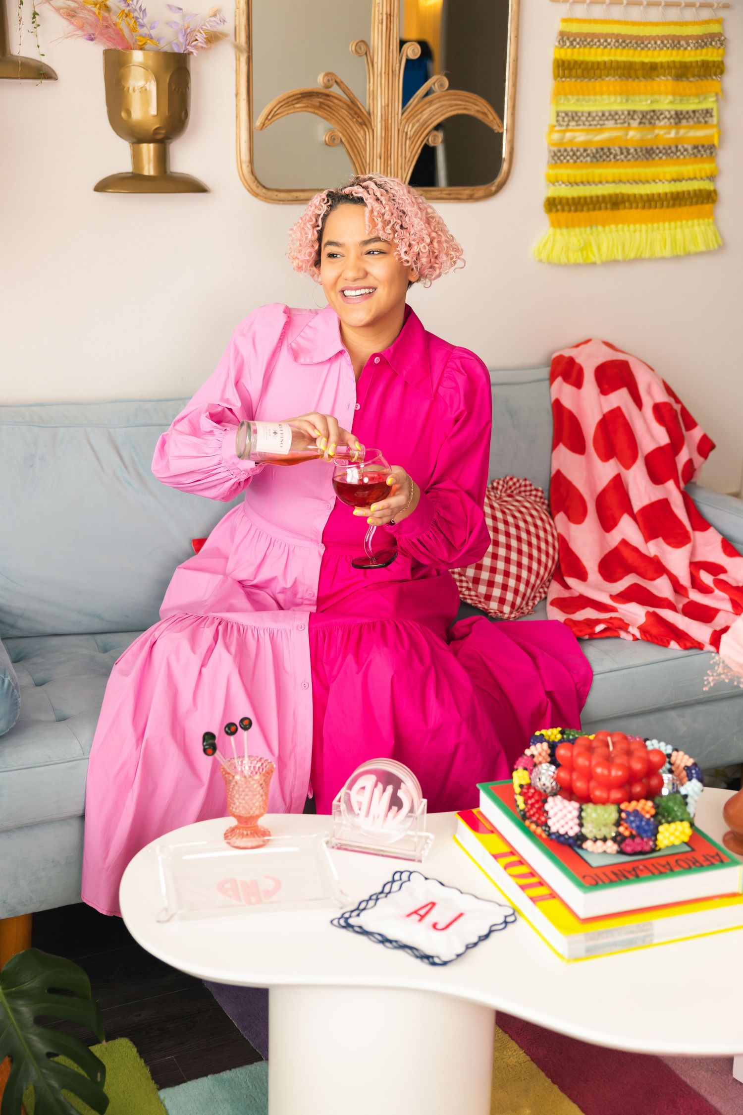 Smiling woman in two-tone pink outfit sitting on blue sofa with monogrammed cake and personalized gifts on white table.