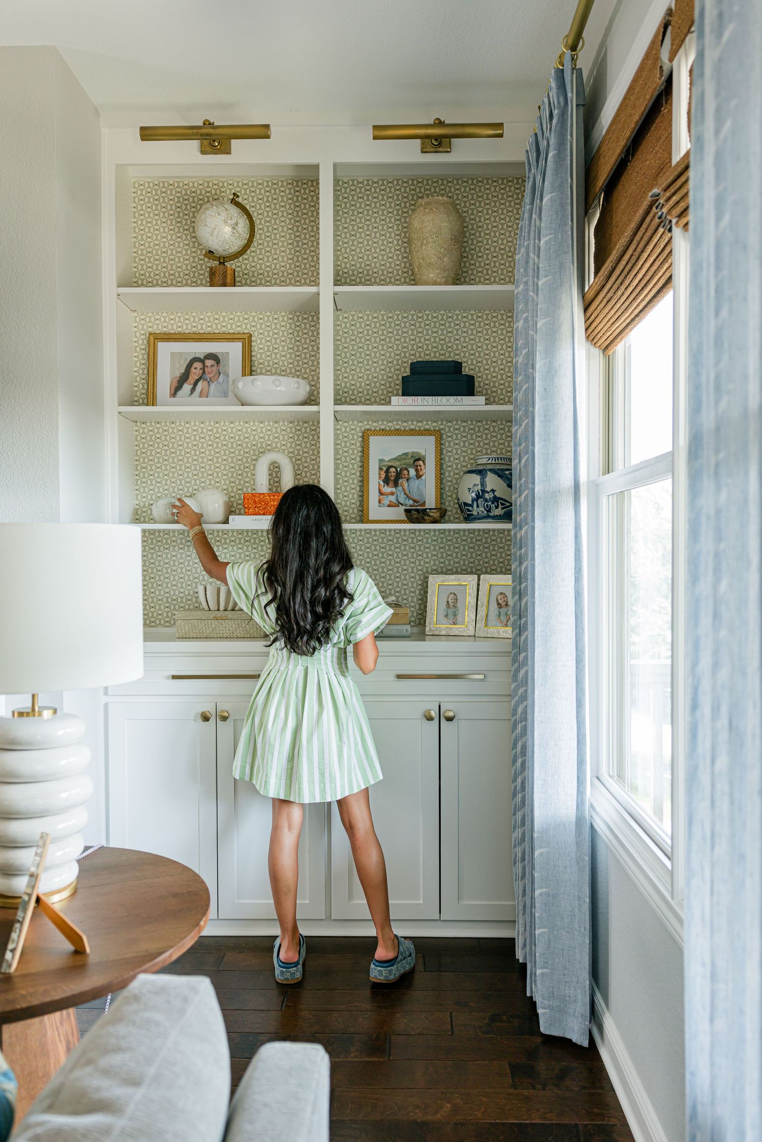 Woman in green dress arranging decor on built-in white shelves with patterned wallpaper backing and brass accents.