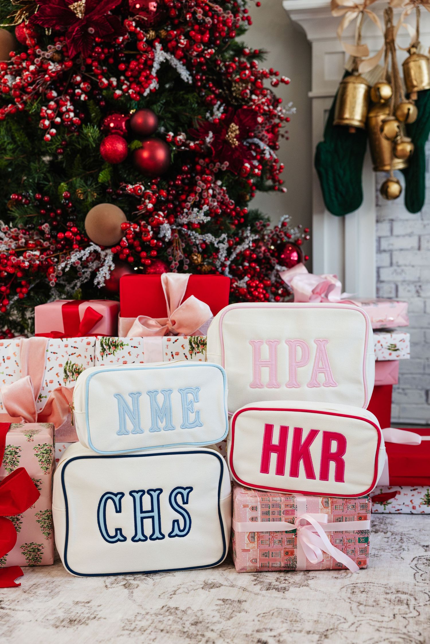 Four monogrammed cosmetic cases arranged as Christmas gifts near holiday tree.