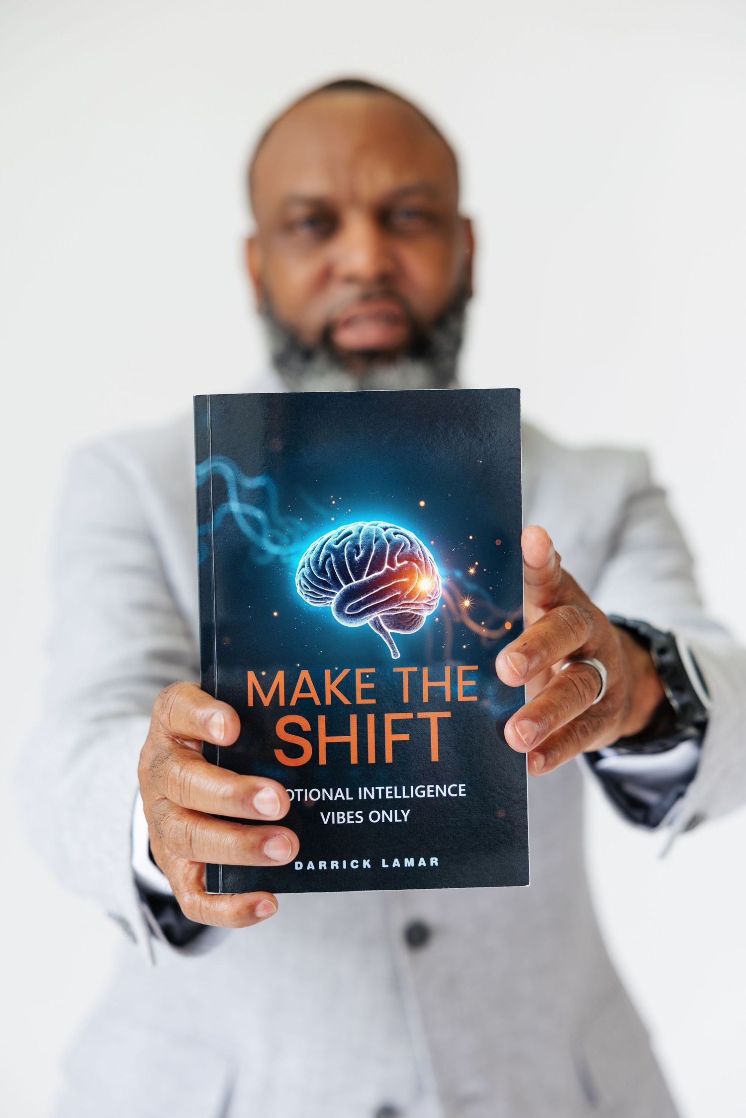 Man in gray blazer holding Make The Shift emotional intelligence book by Darrick Lamar toward camera.