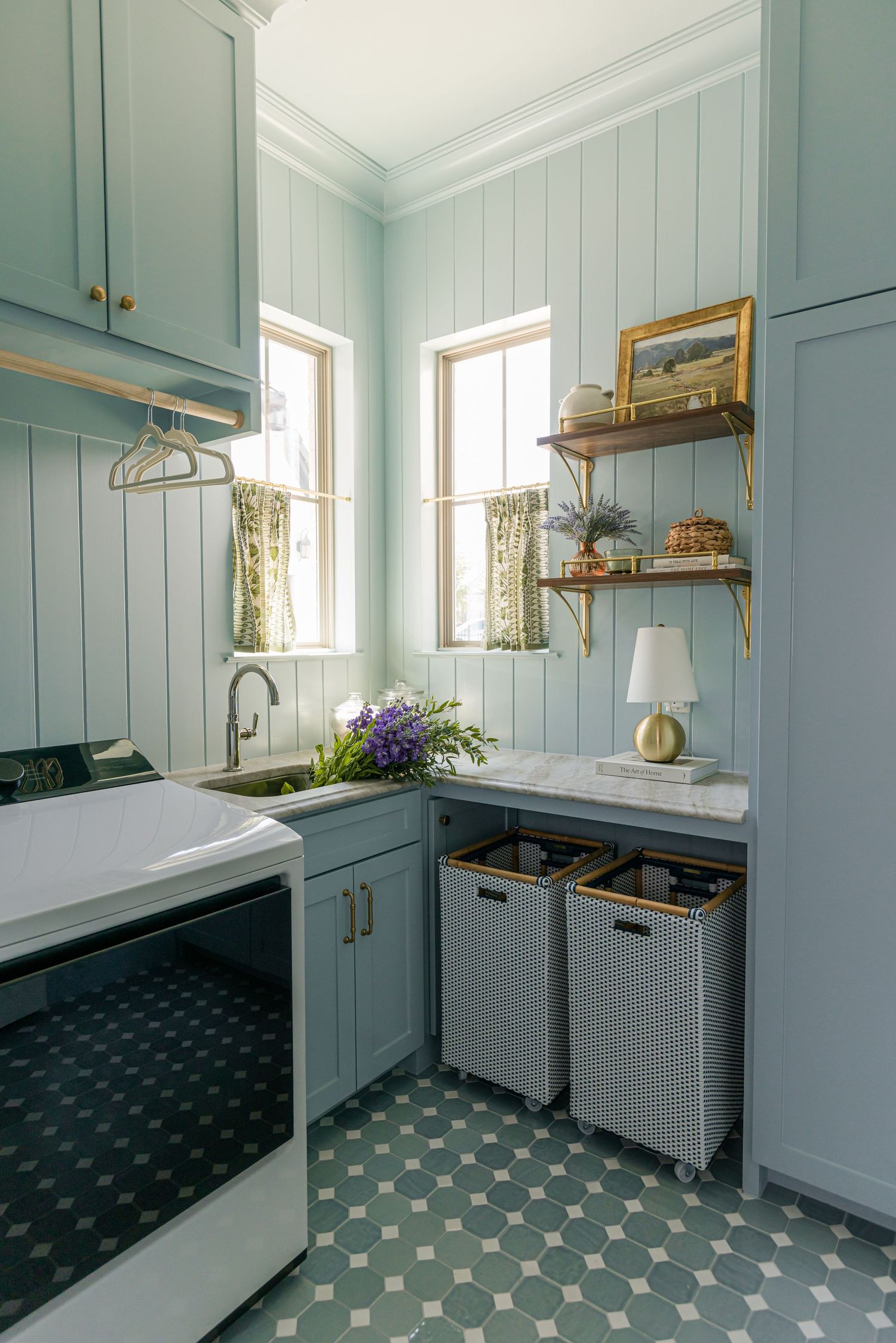 Charming blue laundry room with patterned tile floor, marble counter, open wood shelves, and floral cafe curtains.