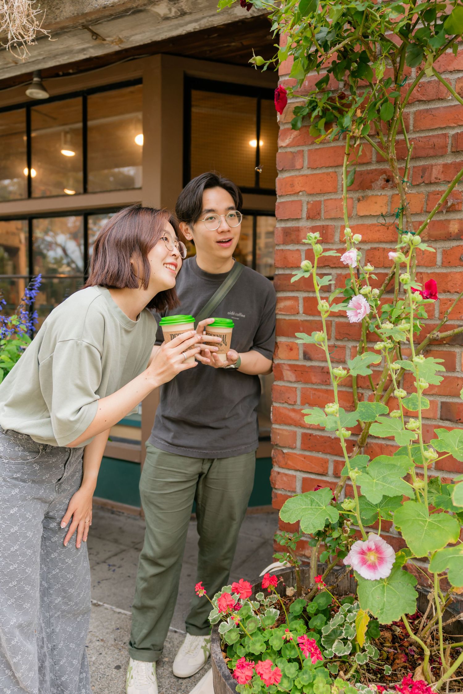 A smiling couple holding green cups outside a brick building surrounded by colorful flowers.