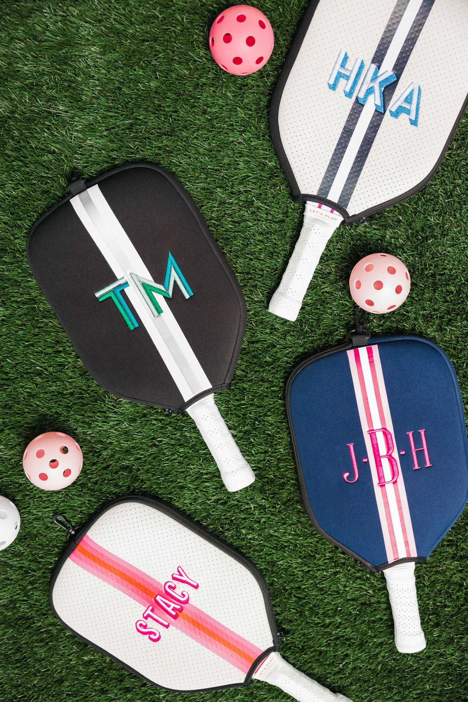 Four monogrammed pickleball paddles in black, white, and navy displayed on green turf with pink wiffle balls.