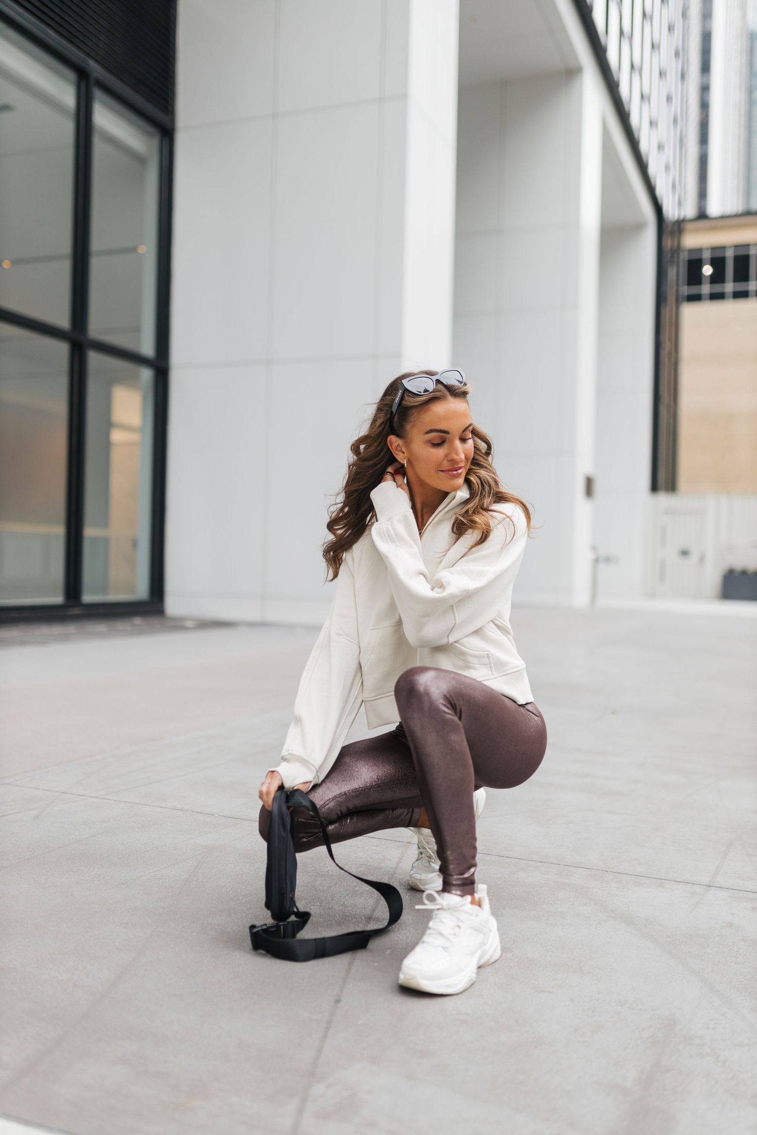 Stylish woman in white jacket and brown metallic leggings crouching outdoors holding a black camera grip.