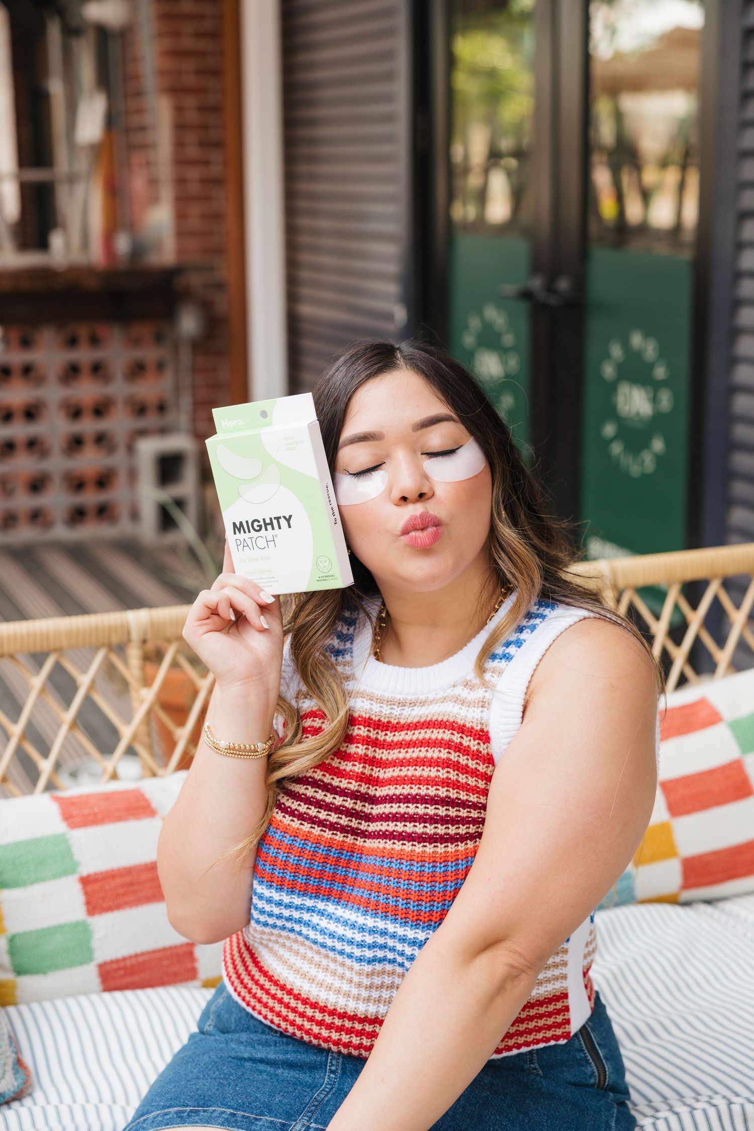 Woman holding Mighty Patch acne patches while sitting outdoors on a colorful striped cushioned chair.