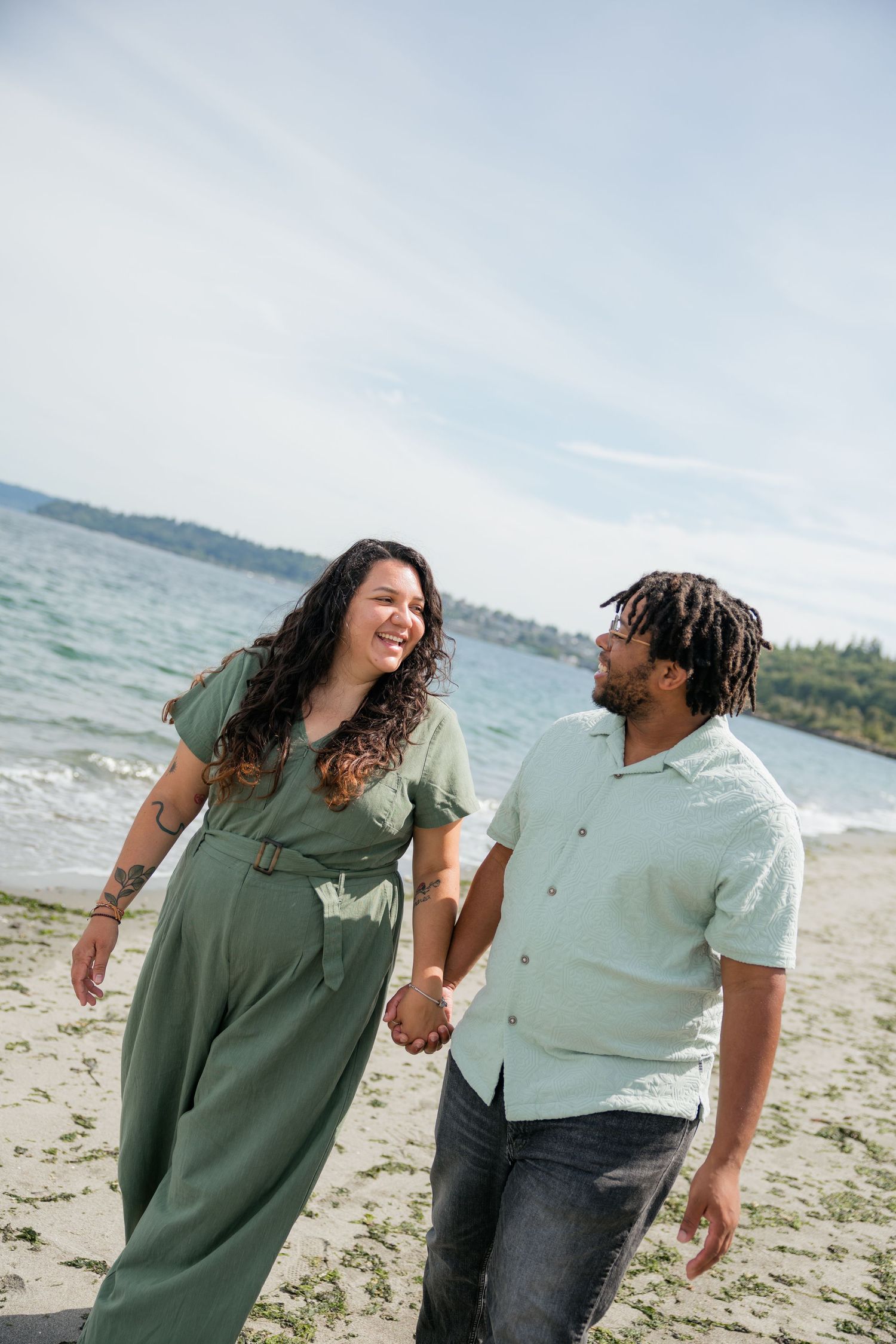 Smiling couple holds hands and gazes at each other while walking along a bright sandy beach.