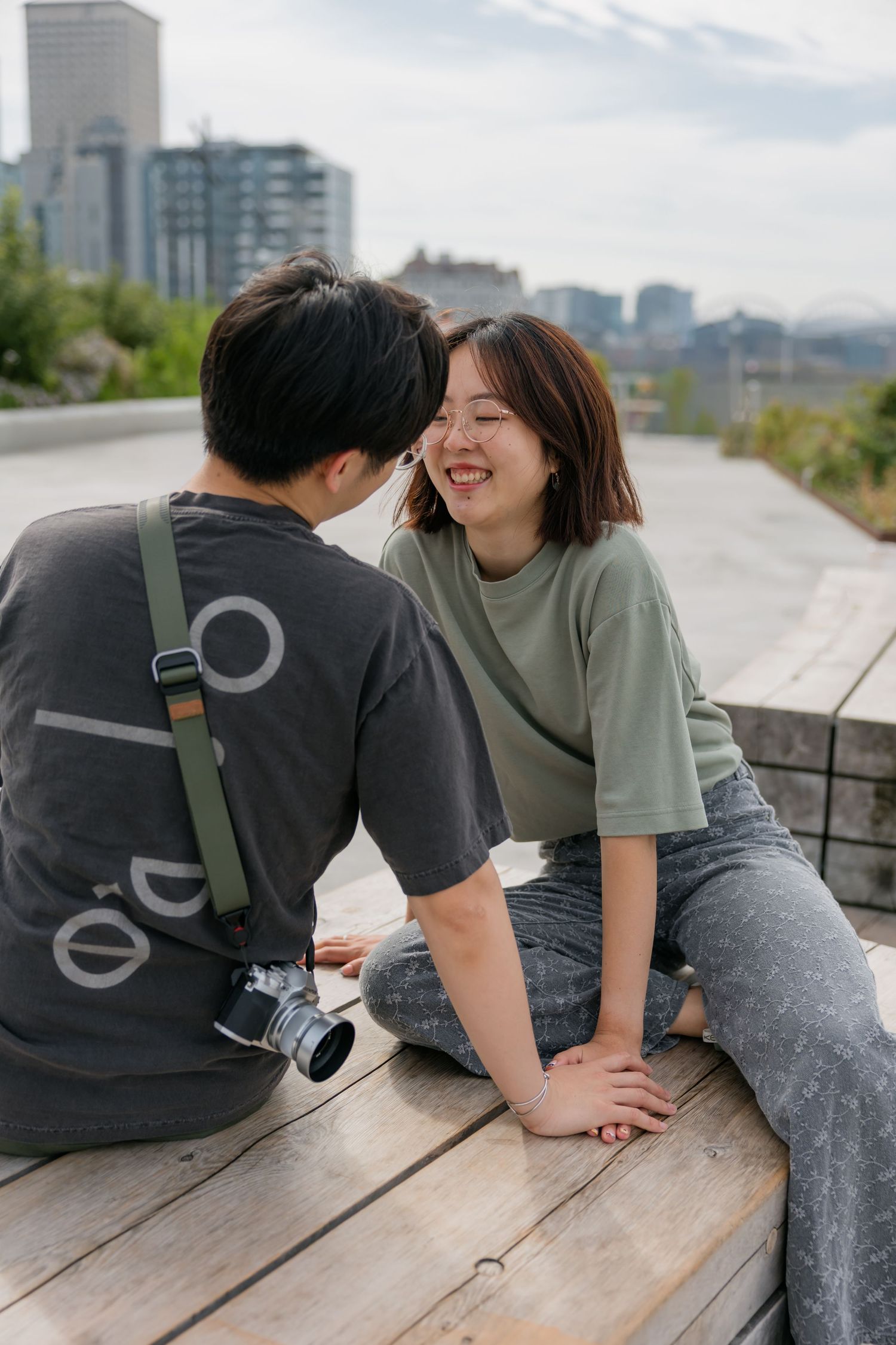 Young couple sitting on wooden steps outdoors, woman smiling joyfully, man with camera on green strap.