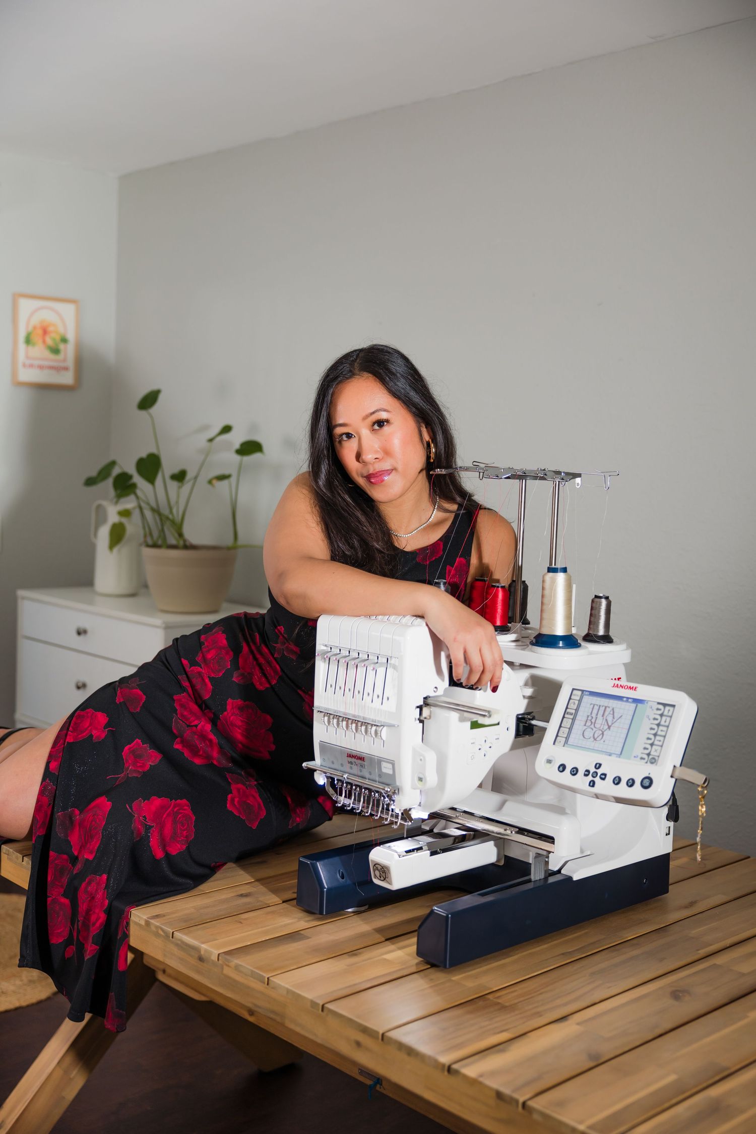 Woman in floral dress posing with an embroidery sewing machine on a wooden table in a bright studio.