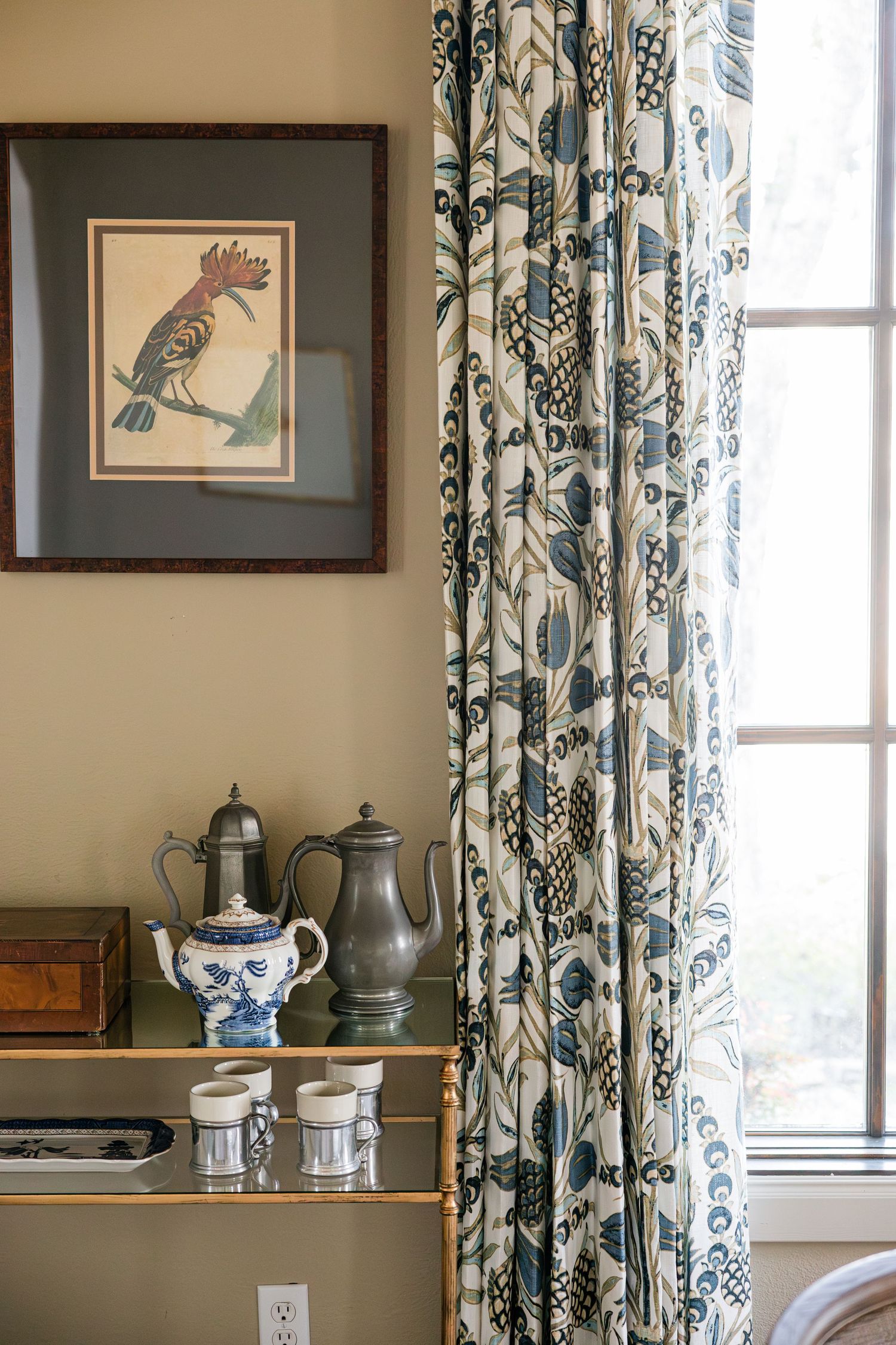 Blue and white floral curtains beside a brass shelf displaying pewter teapots and a framed bird artwork.