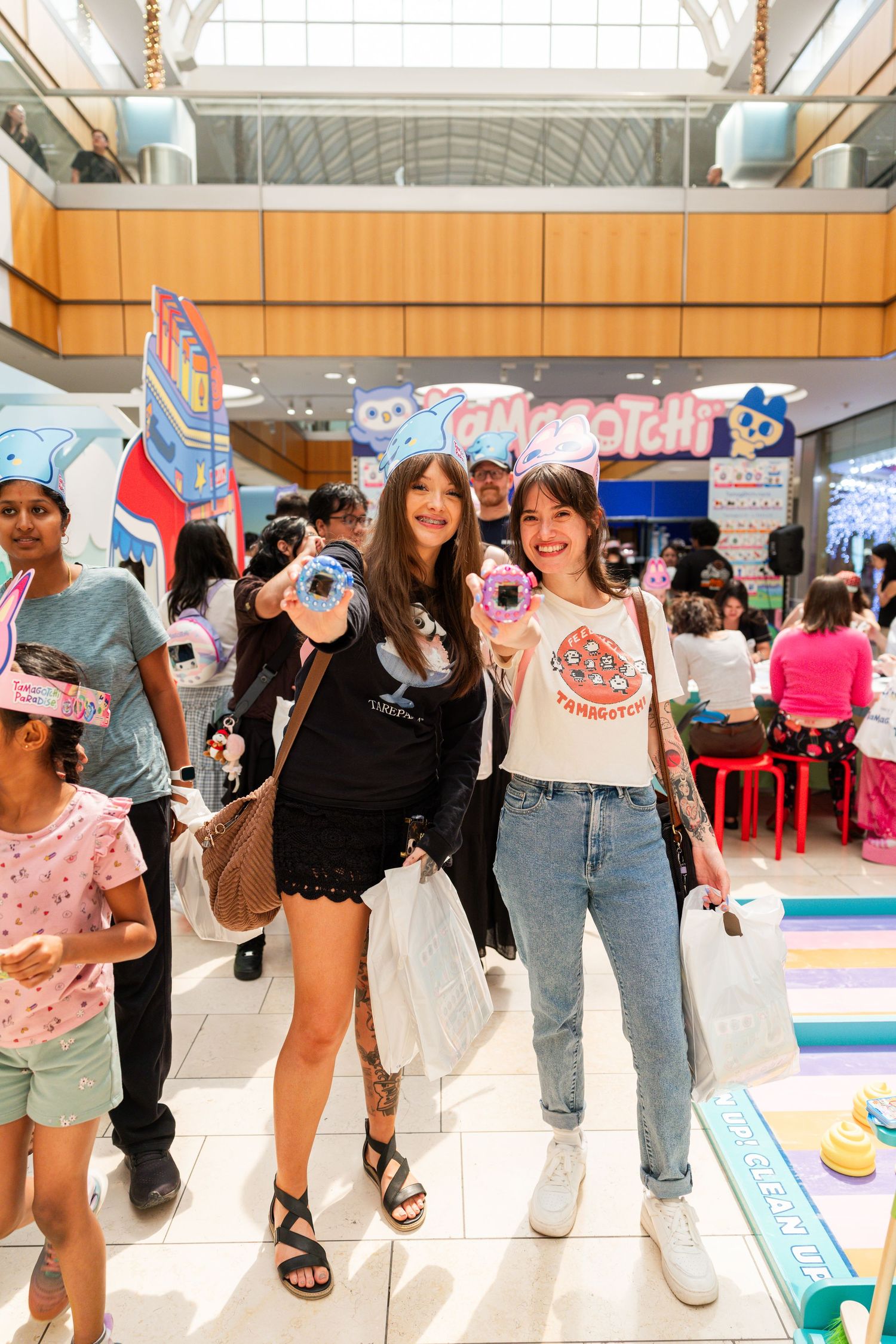 Two women pose with Tamagotchi devices at a colorful Tamagotchi pop-up event in a shopping mall.