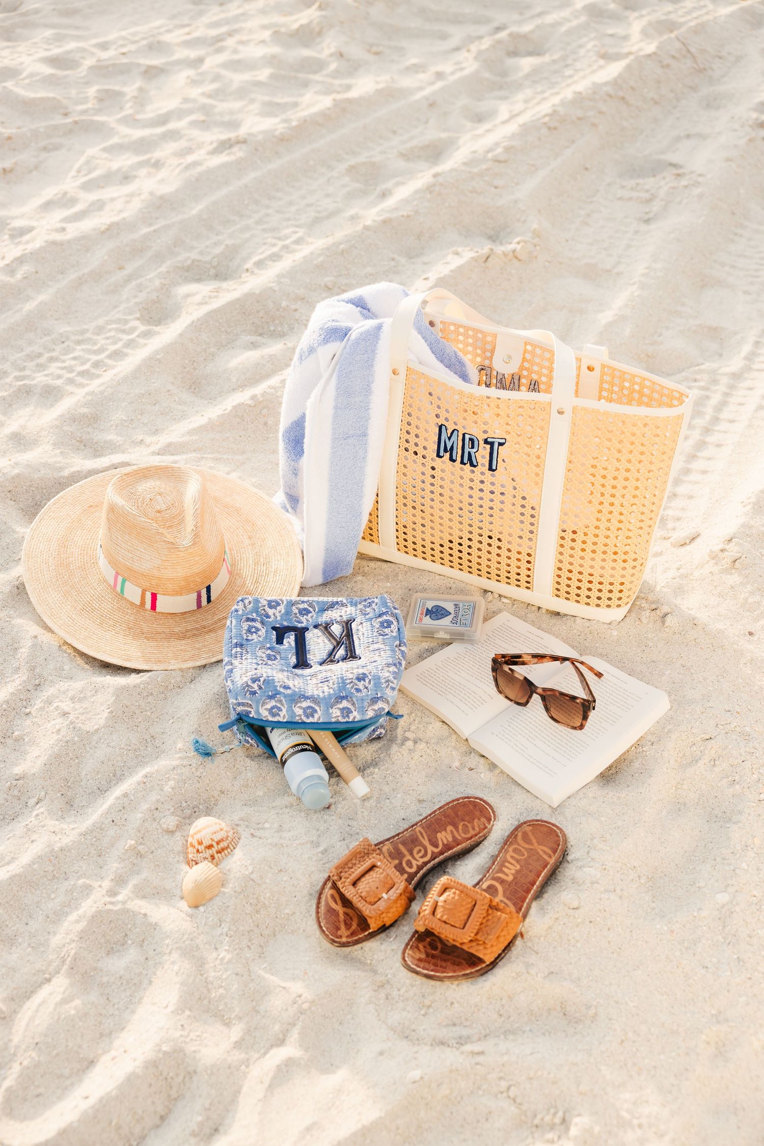 Monogrammed beach tote, straw hat, sandals, sunglasses, and accessories arranged on white sand.
