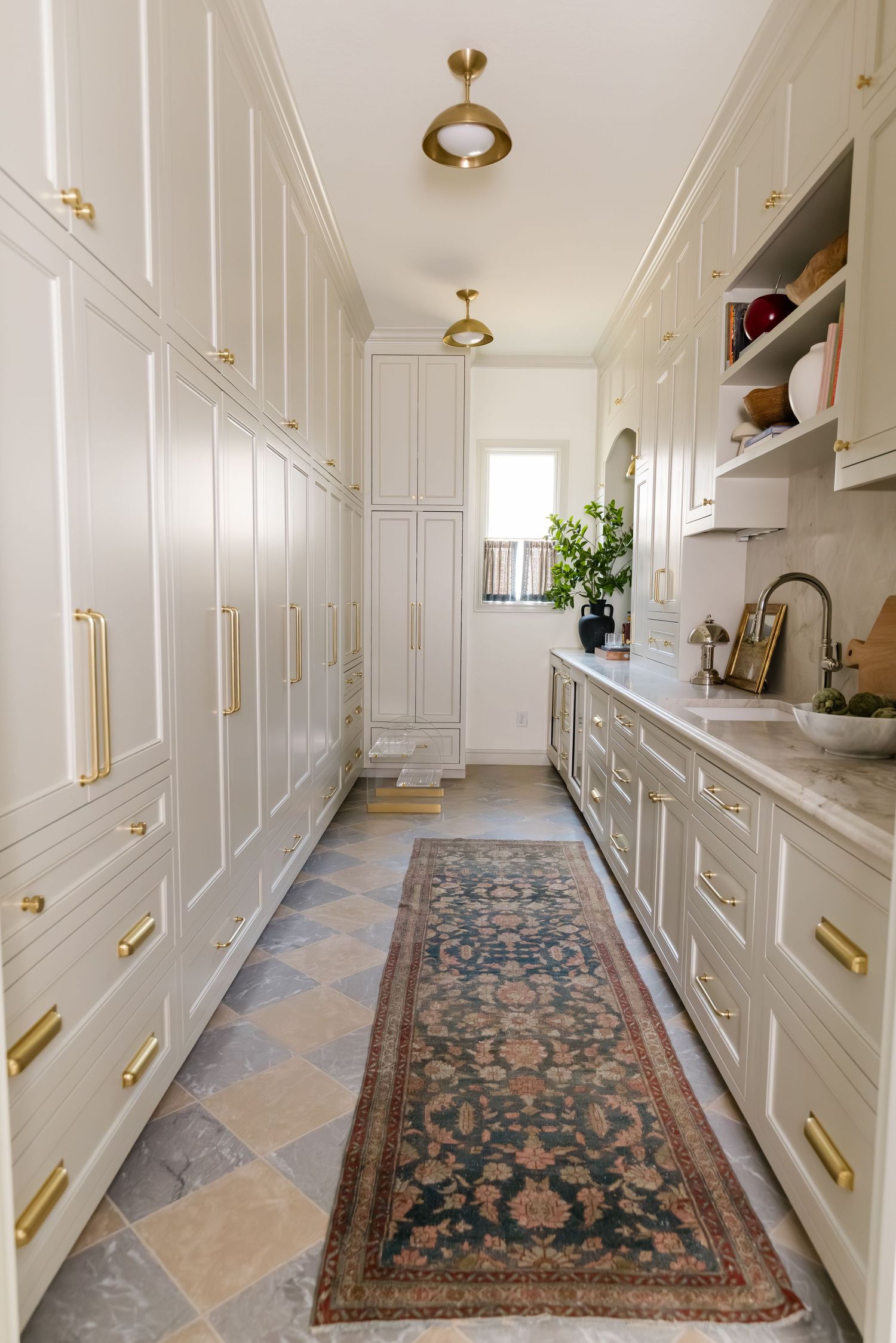 Narrow butler pantry with cream cabinets, brass hardware, stone tile floor, vintage runner rug, and pendant lights.