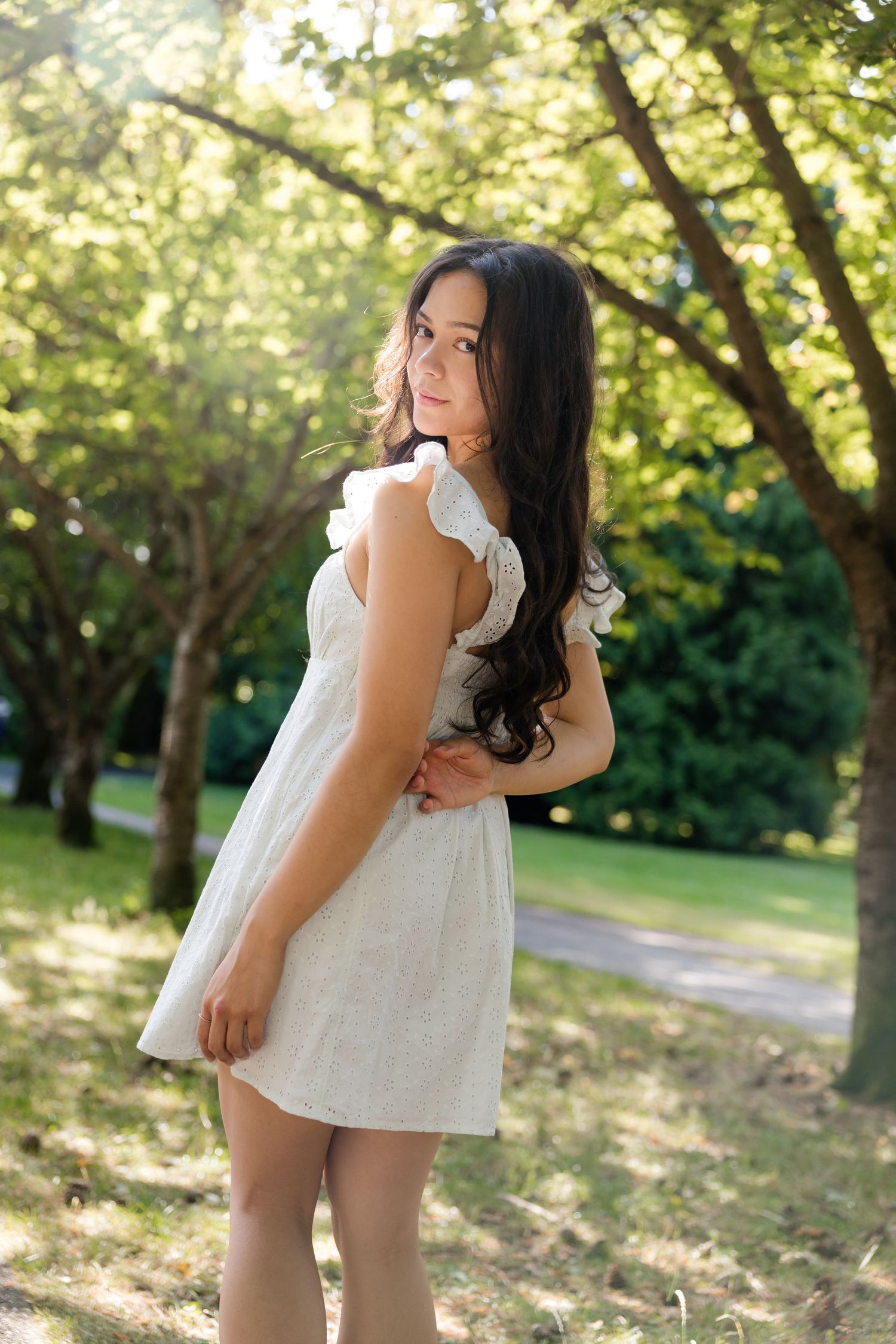 Young woman in white ruffled mini dress stands gracefully among sunlit trees in a lush green park.