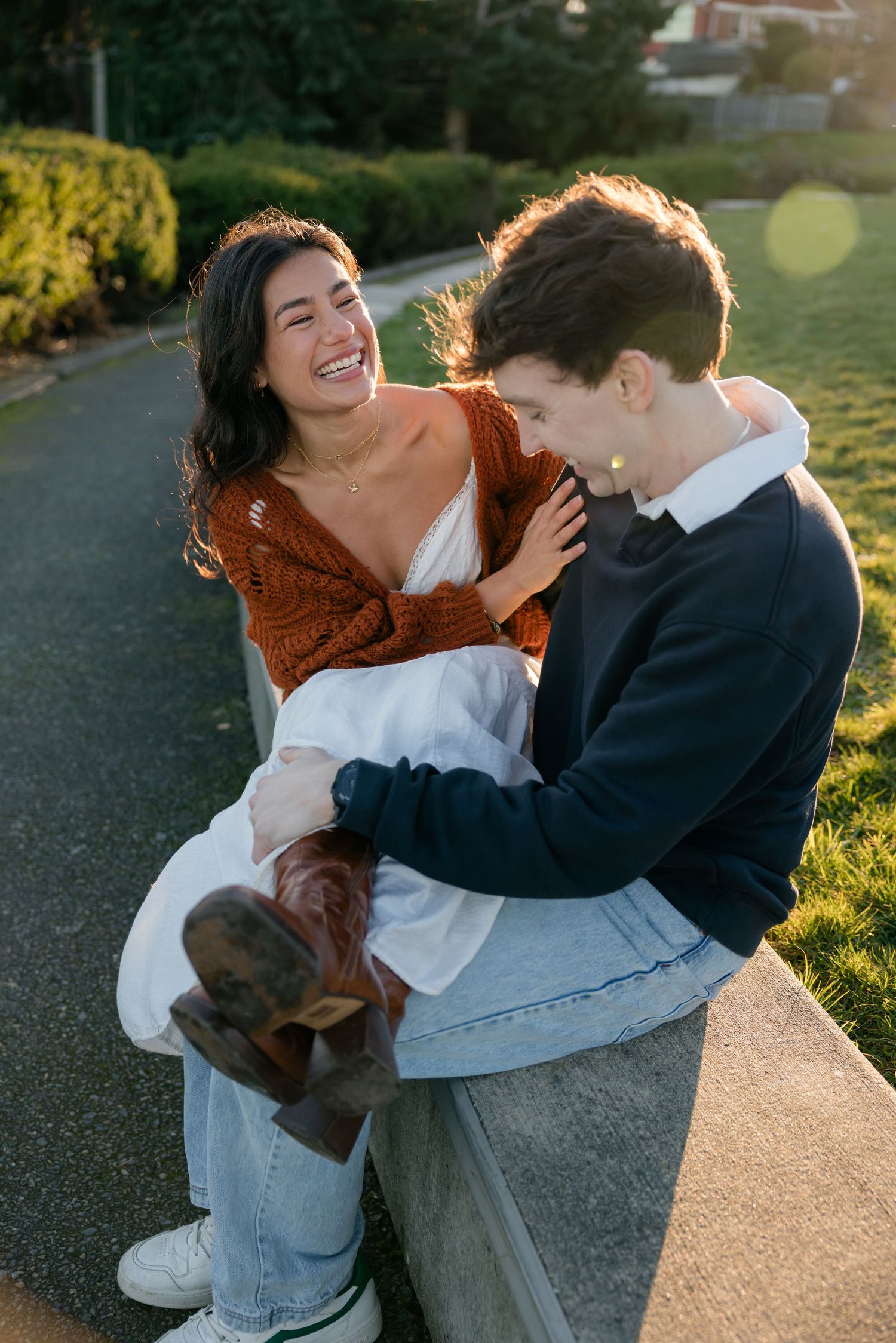 A laughing couple sits together on a ledge in golden sunlight in a lush green park.