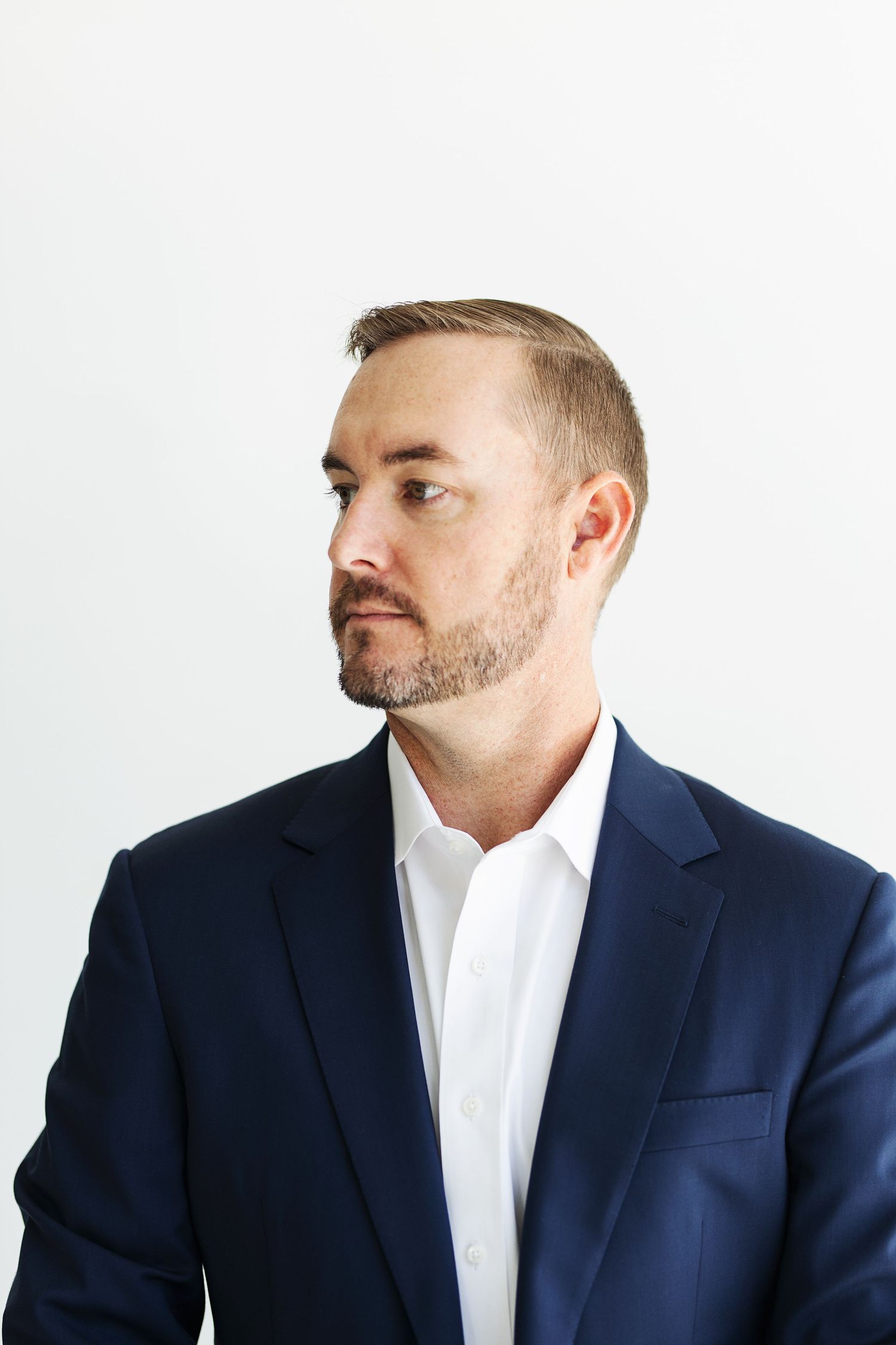 Professional man in navy blazer and white shirt with short beard looking sideways against white background.