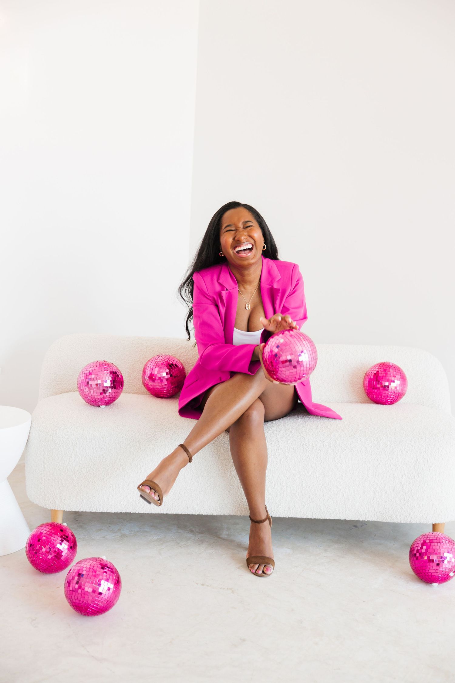 Laughing woman in hot pink blazer sitting on white sofa surrounded by pink disco balls.
