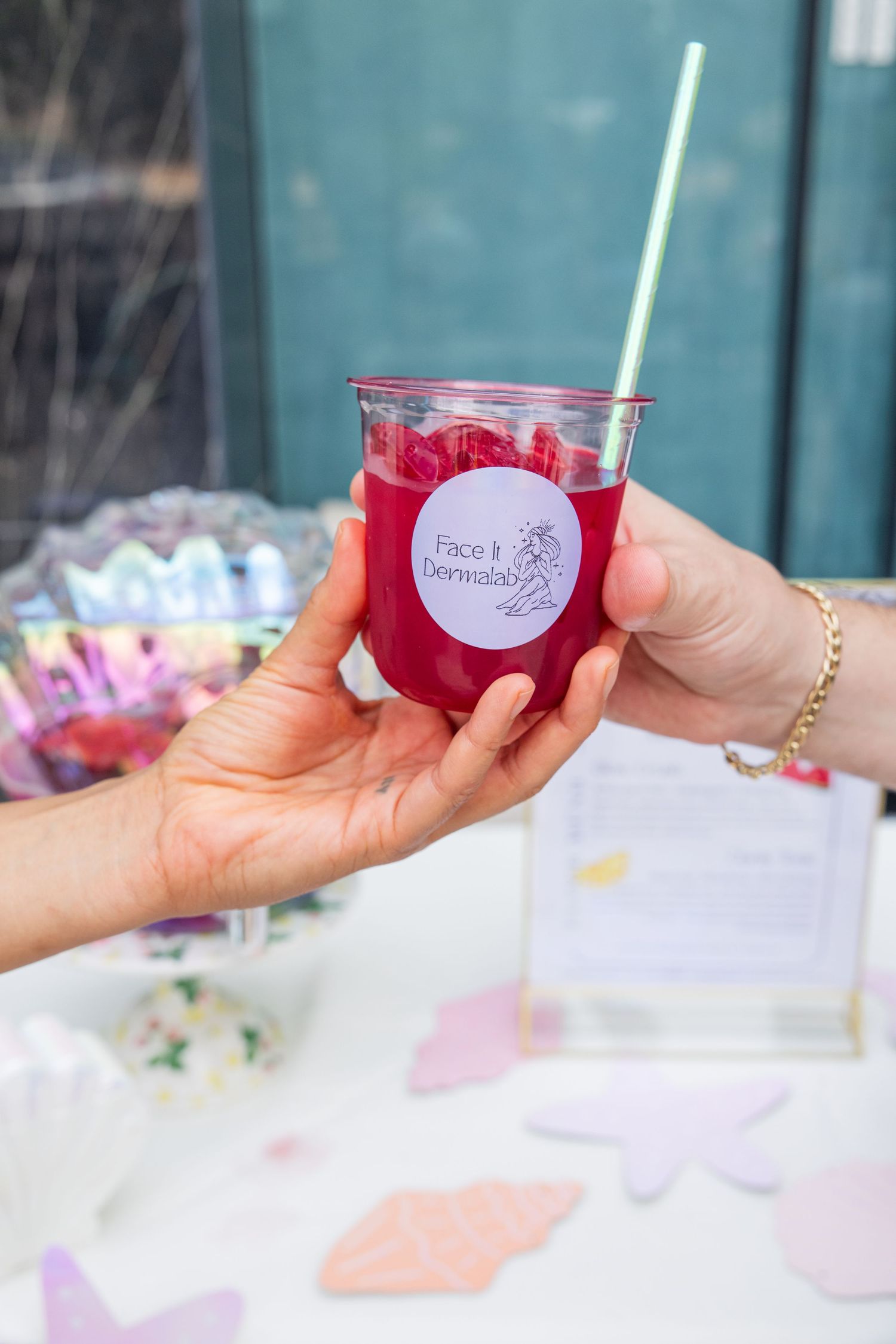 Hand passing a red drink in a branded cup at a Face It Dermatology event.
