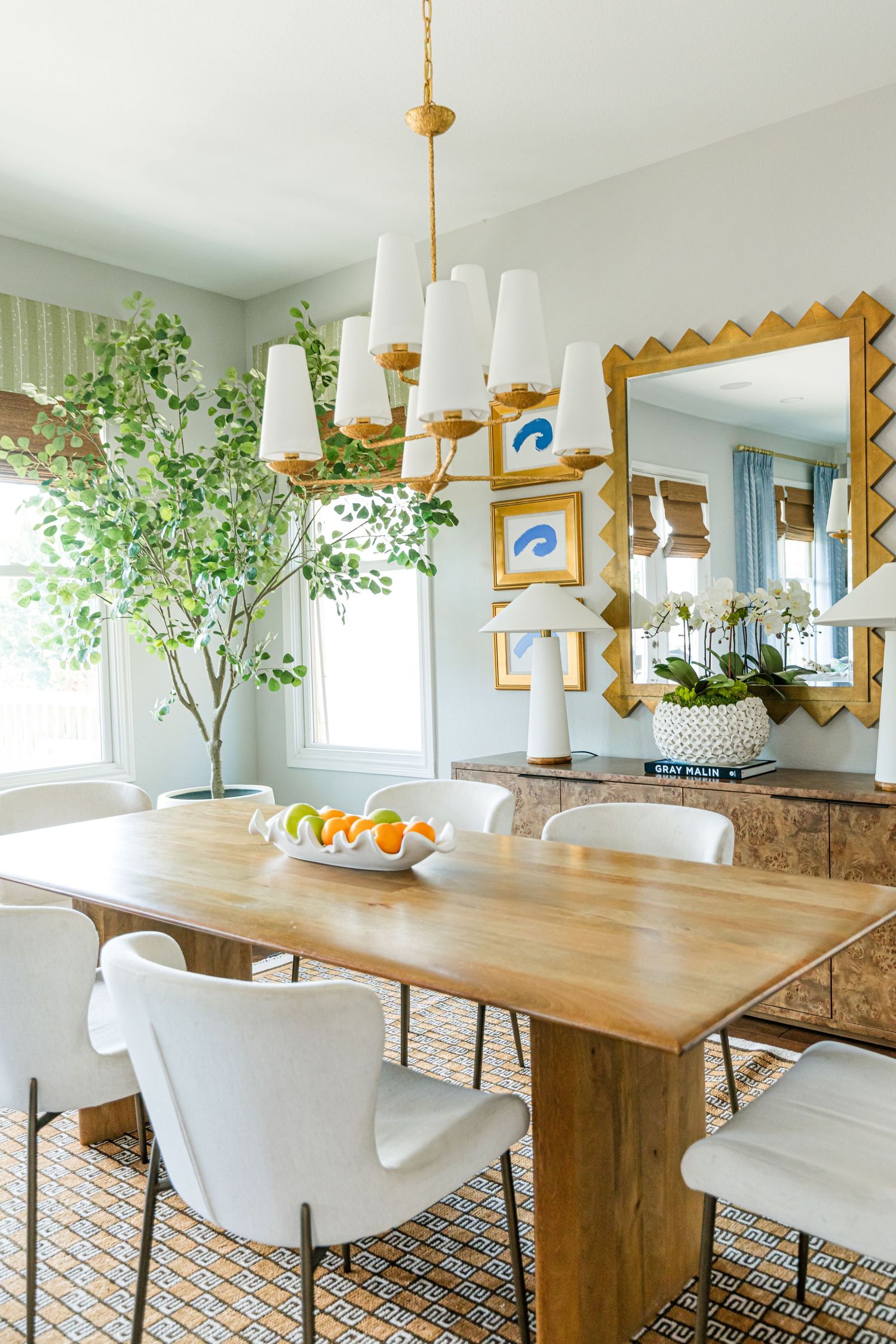 Bright dining room with wood table, white chairs, gold chandelier, zigzag mirror, and tall indoor tree.