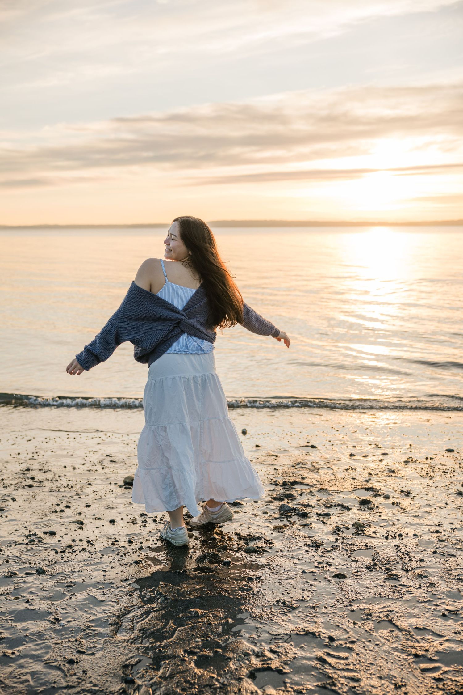 Woman in white dress and blue jacket twirls joyfully on a beach during a stunning golden sunset.