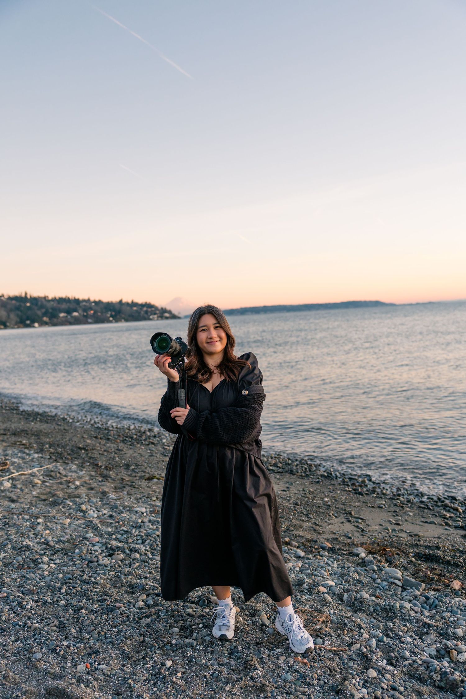 A smiling woman in a black dress holds a camera on a rocky beach at sunset.