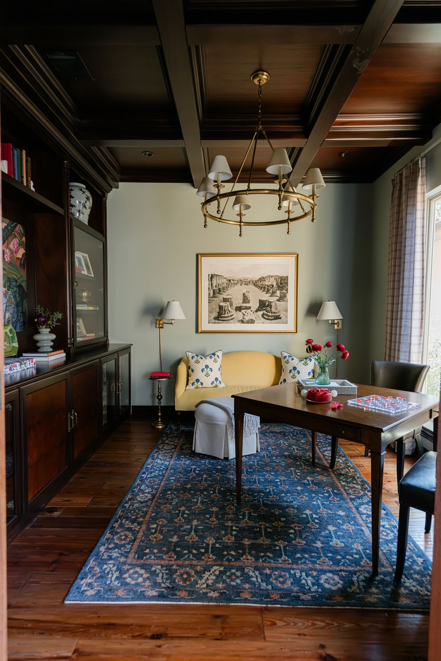 Sophisticated study with dark coffered ceiling, blue oriental rug, yellow sofa, brass chandelier, and classic artwork.
